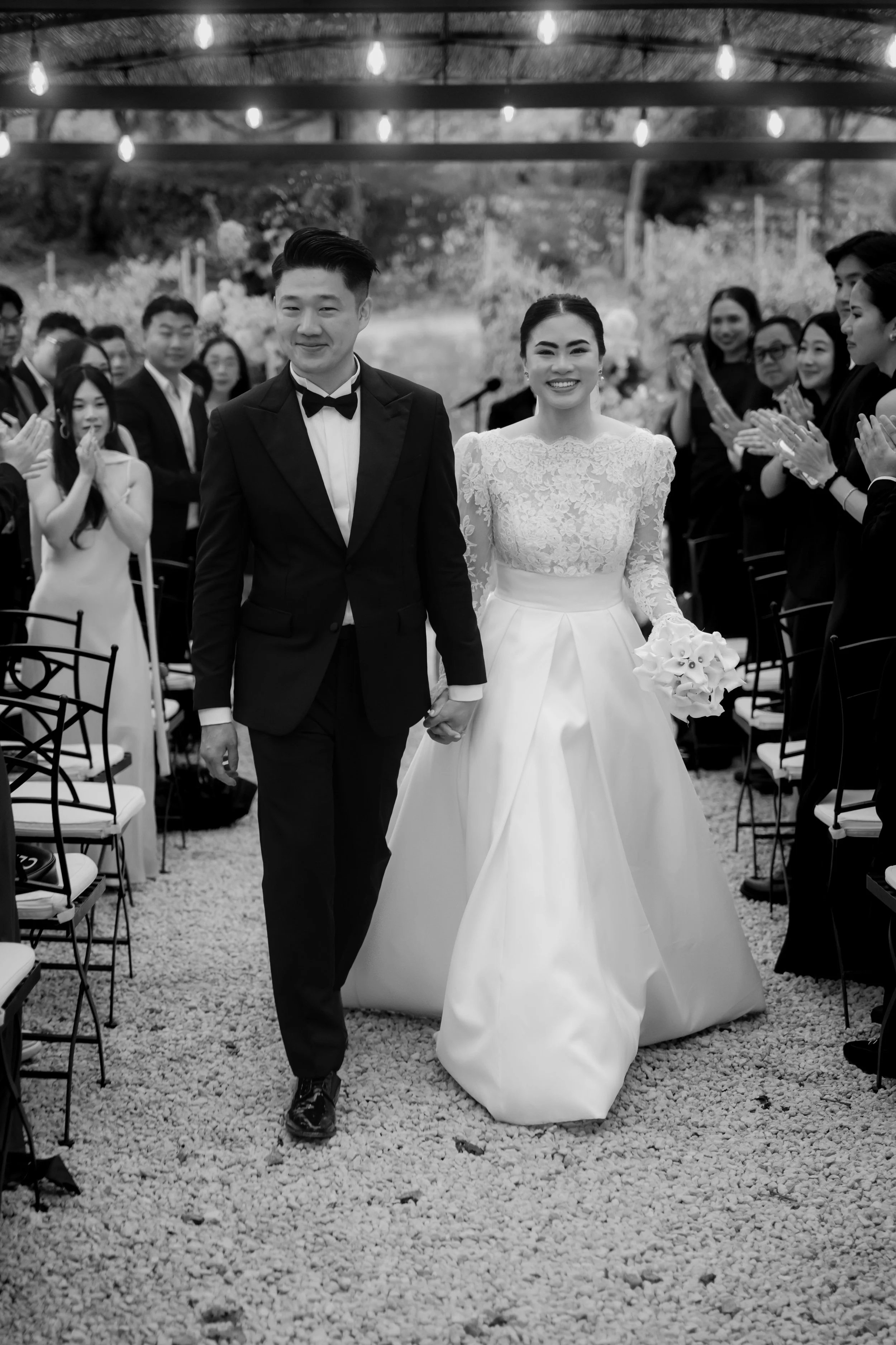 A bride and groom walking hand in hand down the aisle during their wedding ceremony, surrounded by seated guests clapping and smiling.