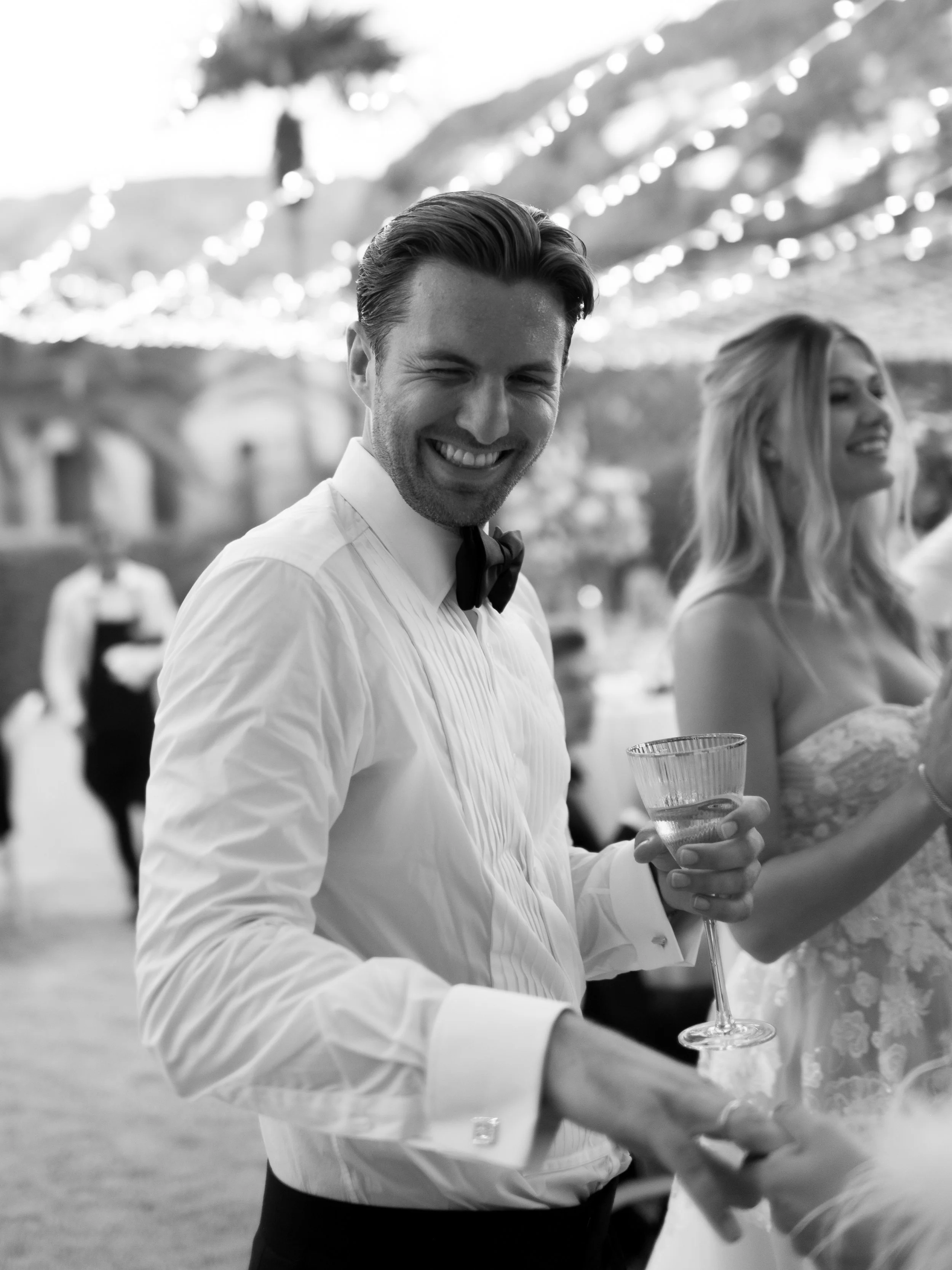 A smiling man in a tuxedo holding a cocktail at a festive outdoor event at dusk, with string lights overhead and a woman in a strapless dress in the background.