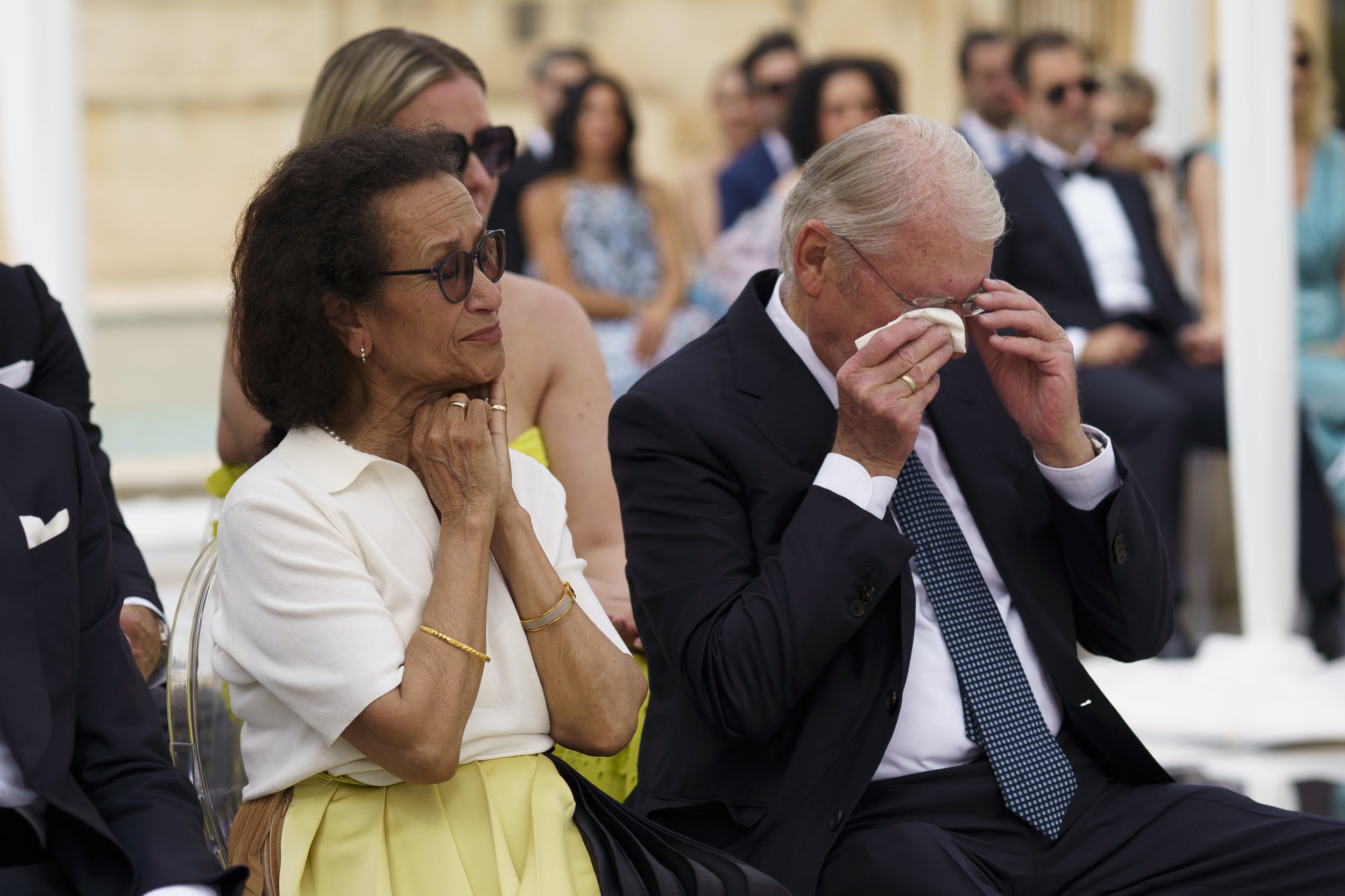 An emotional moment at a formal outdoor event where an elderly man and woman are seated, with the man wiping tears from his eyes, and the woman appearing teary-eyed.