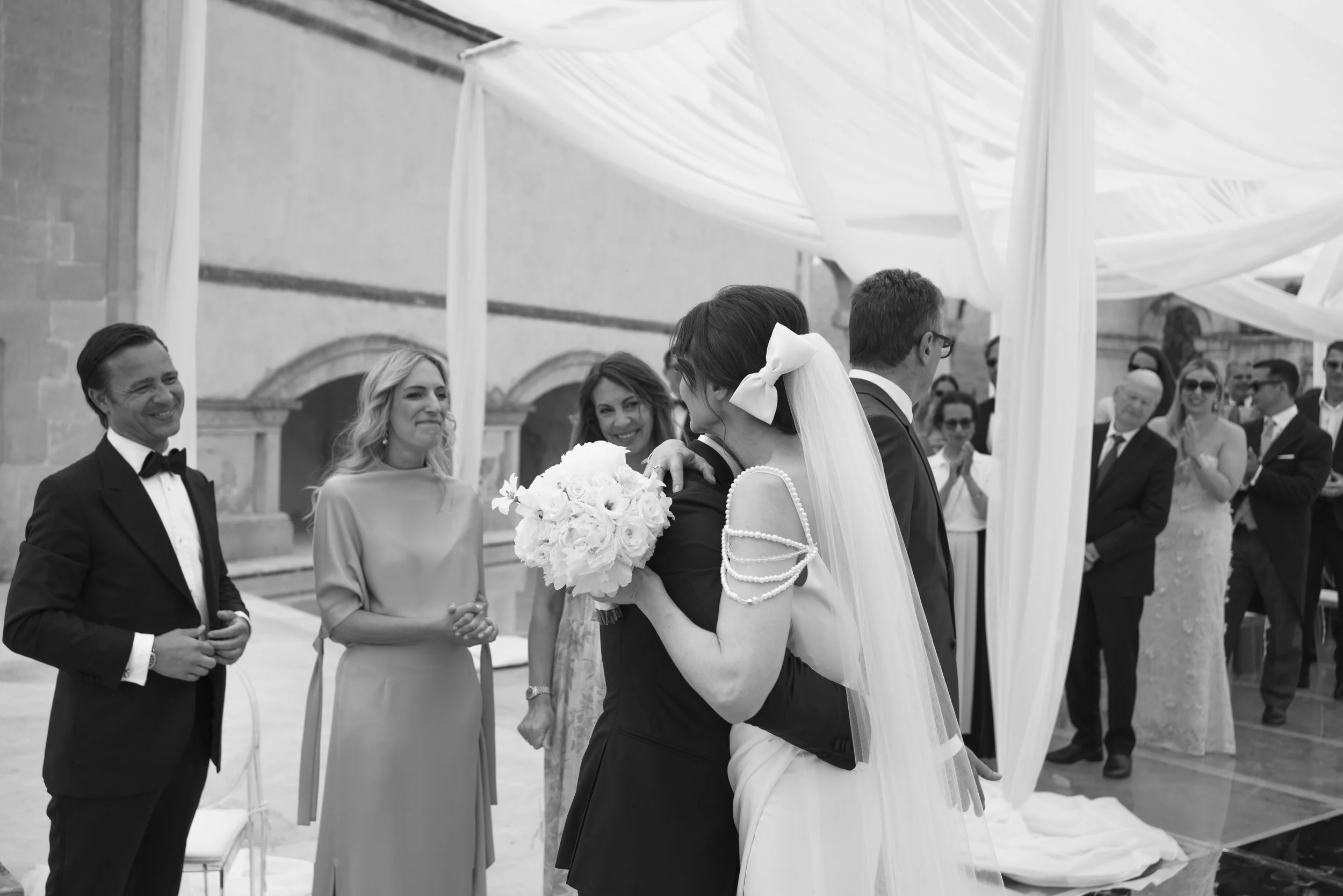A black and white photo of a wedding ceremony with a bride hugging a woman holding a bouquet, surrounded by guests in formal attire.