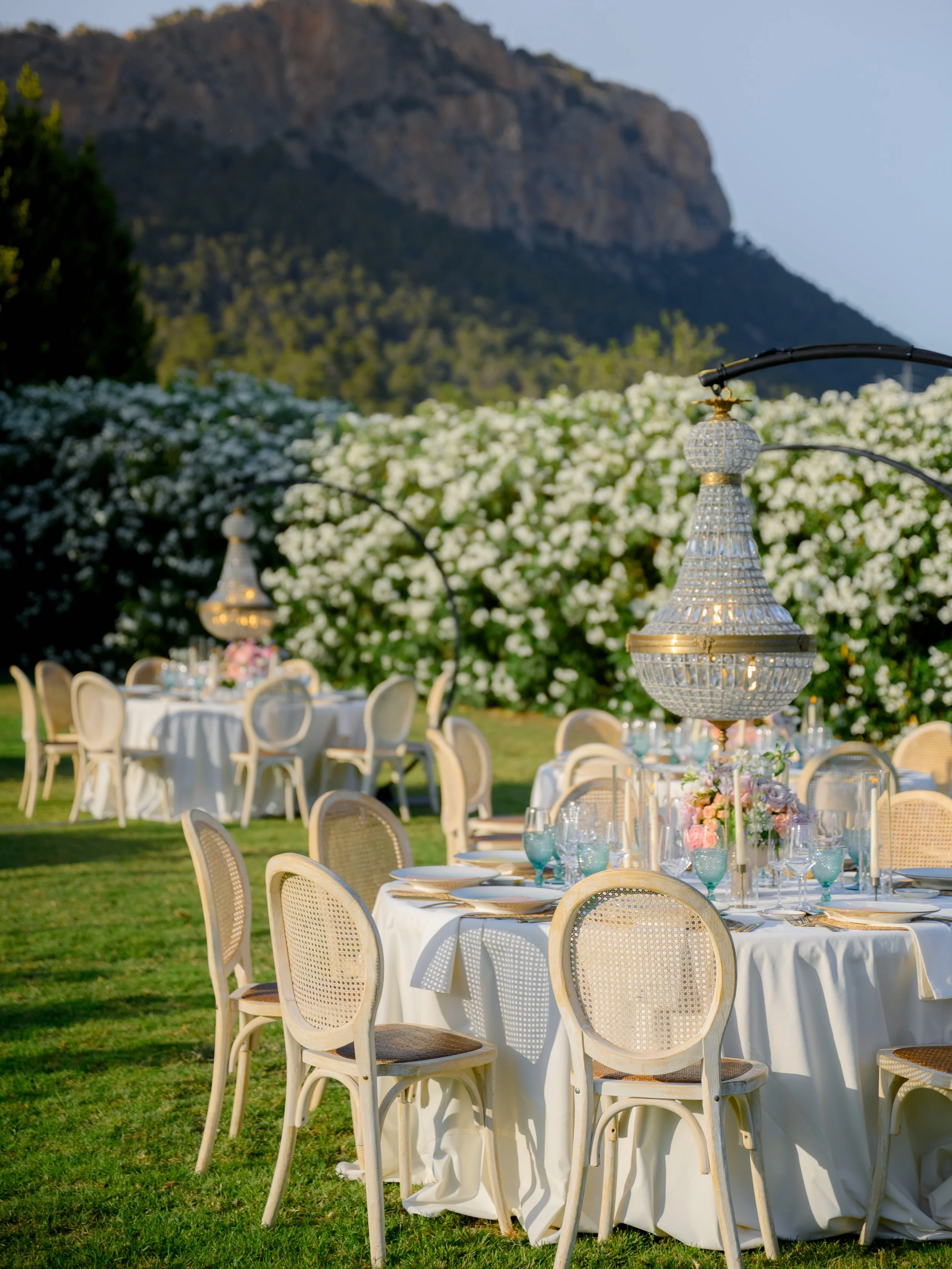 Elegant outdoor dining setup with round tables covered in white tablecloths, surrounded by beige chairs with cane backs. The tables are set with plates, glasses, and floral centerpieces, and a large, ornate crystal chandelier hangs above. In the back