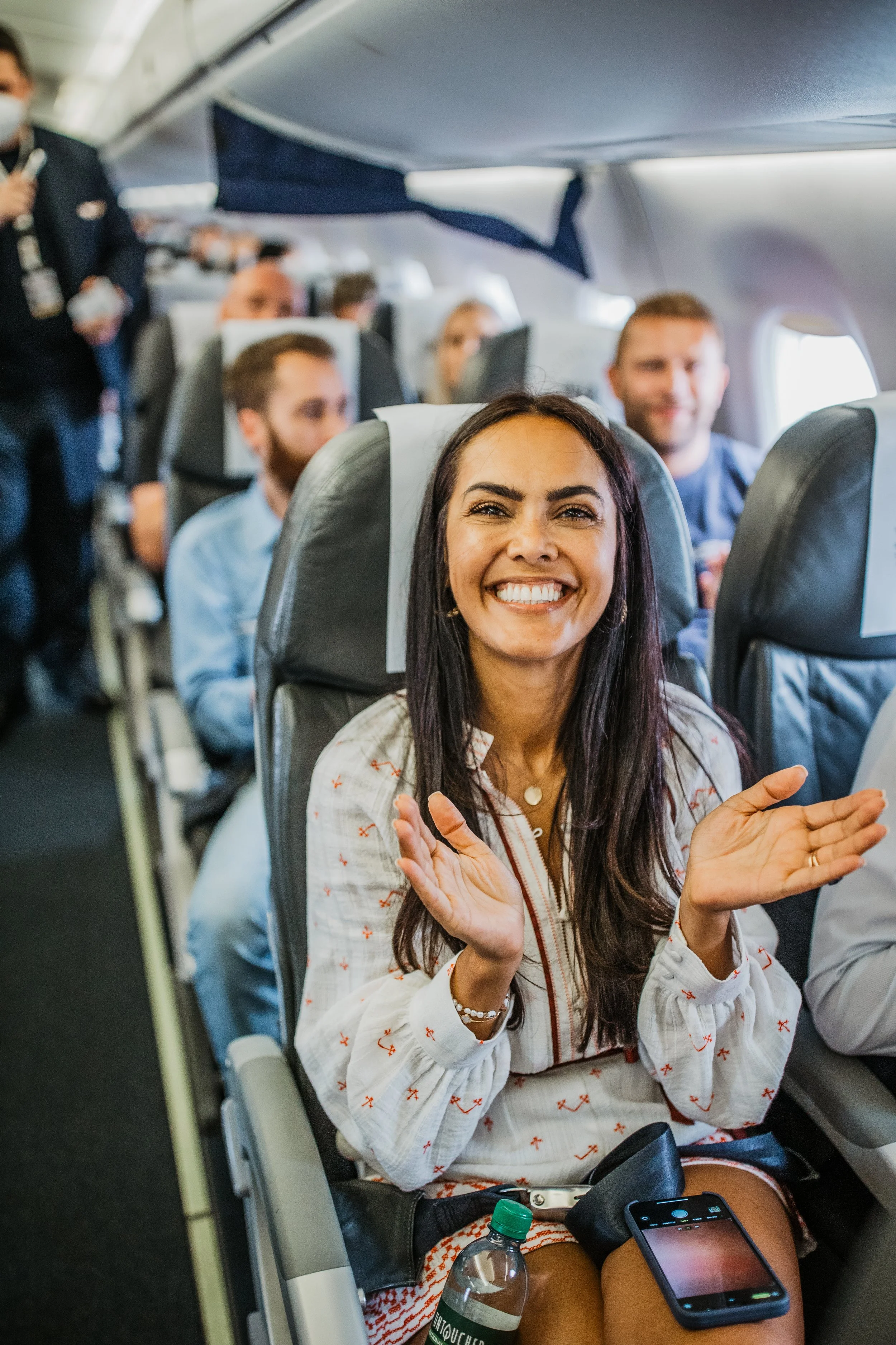 A woman smiling and clapping in an airplane seat, with other passengers and flight attendants visible in the background.