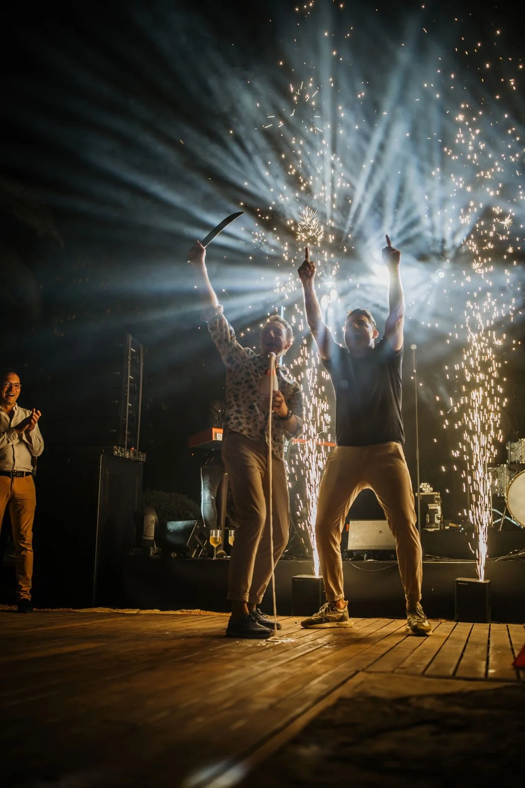 Two men on a stage celebrating with sparklers and fireworks, one holding a knife, at a party or event, with other people clapping in the background.