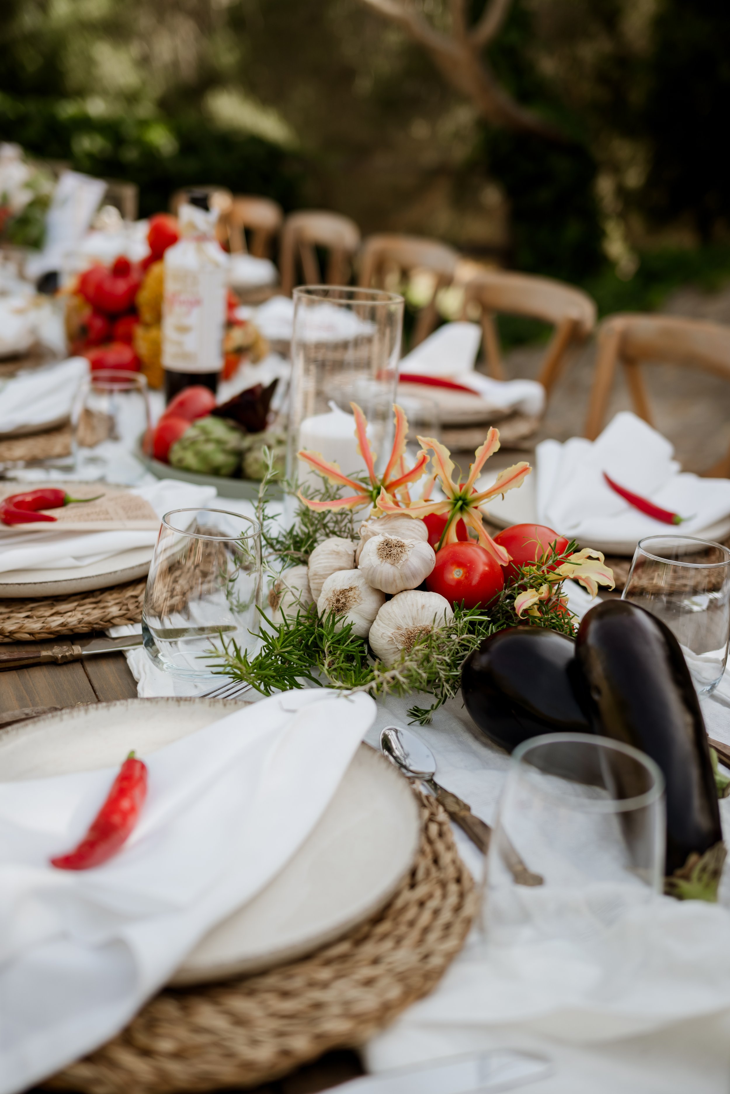 Outdoor dining table with a rustic centerpiece of garlic, tomatoes, eggplants, and flowers, set with plates, glasses, and white napkins.