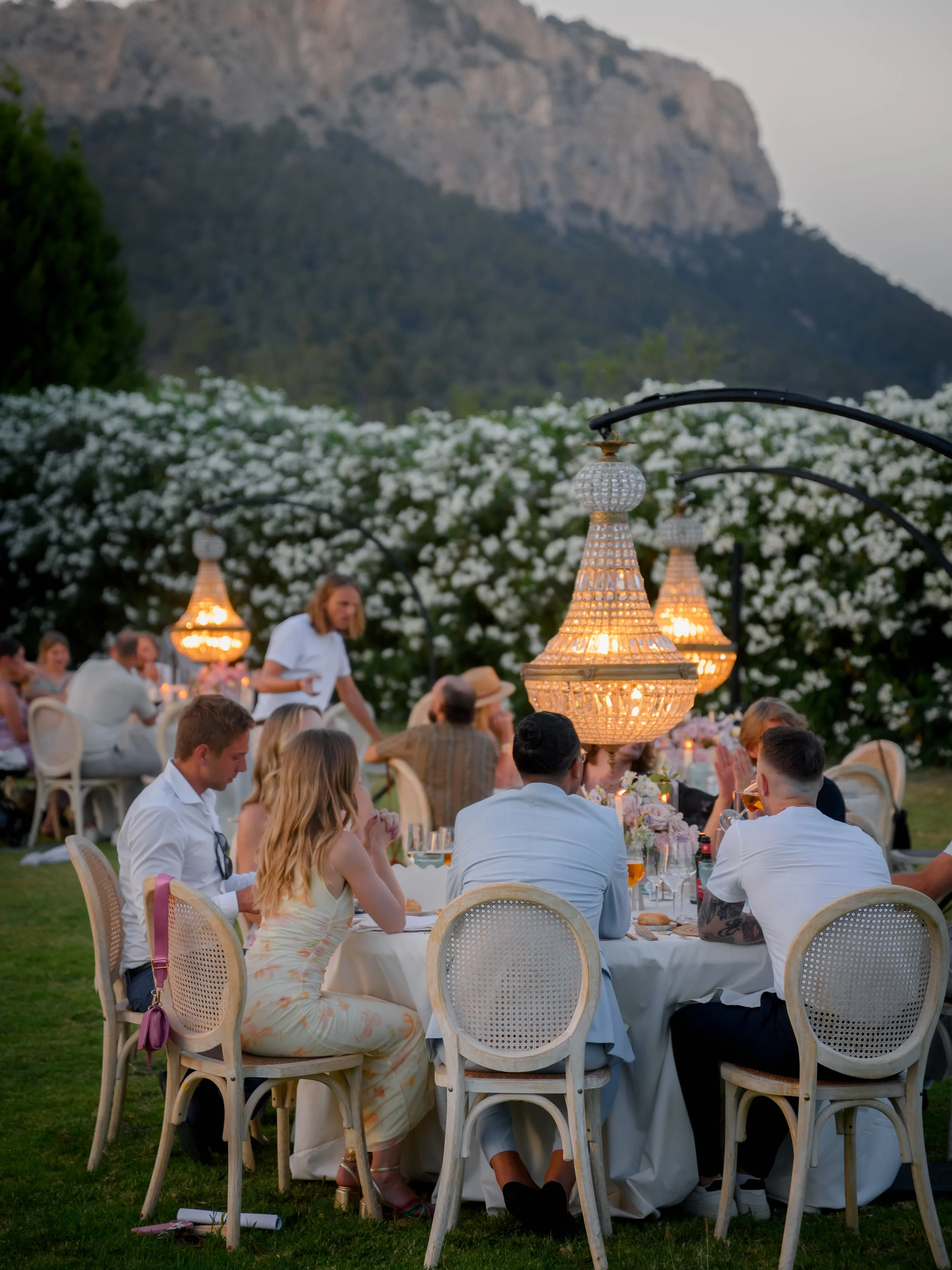 People enjoying a dinner party outdoors at dusk, with large chandeliers hanging over a round table. The setting is in a garden with white flowers and mountains in the background.