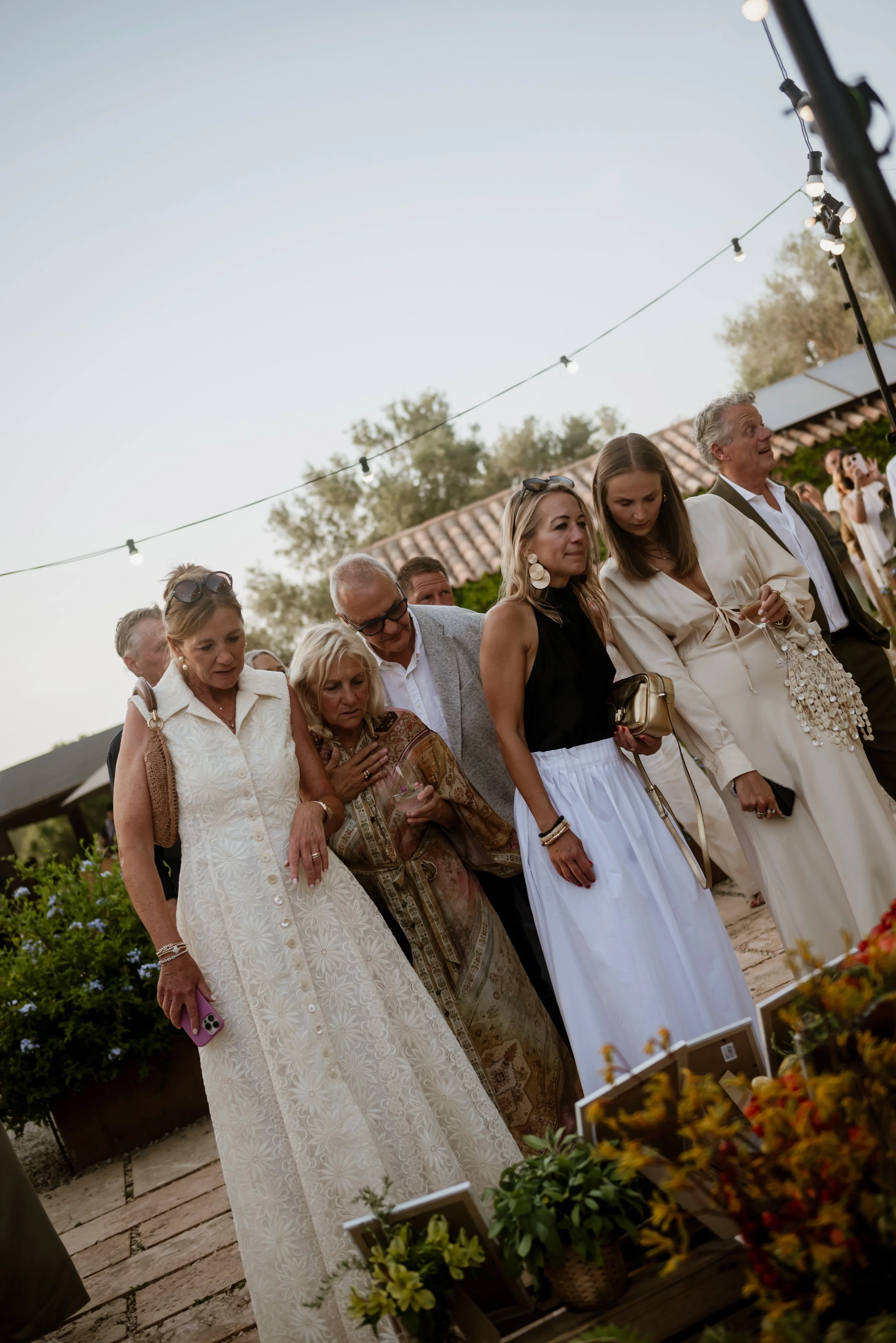 Group of people standing outdoors at a social event, with string lights overhead and plants in the background.