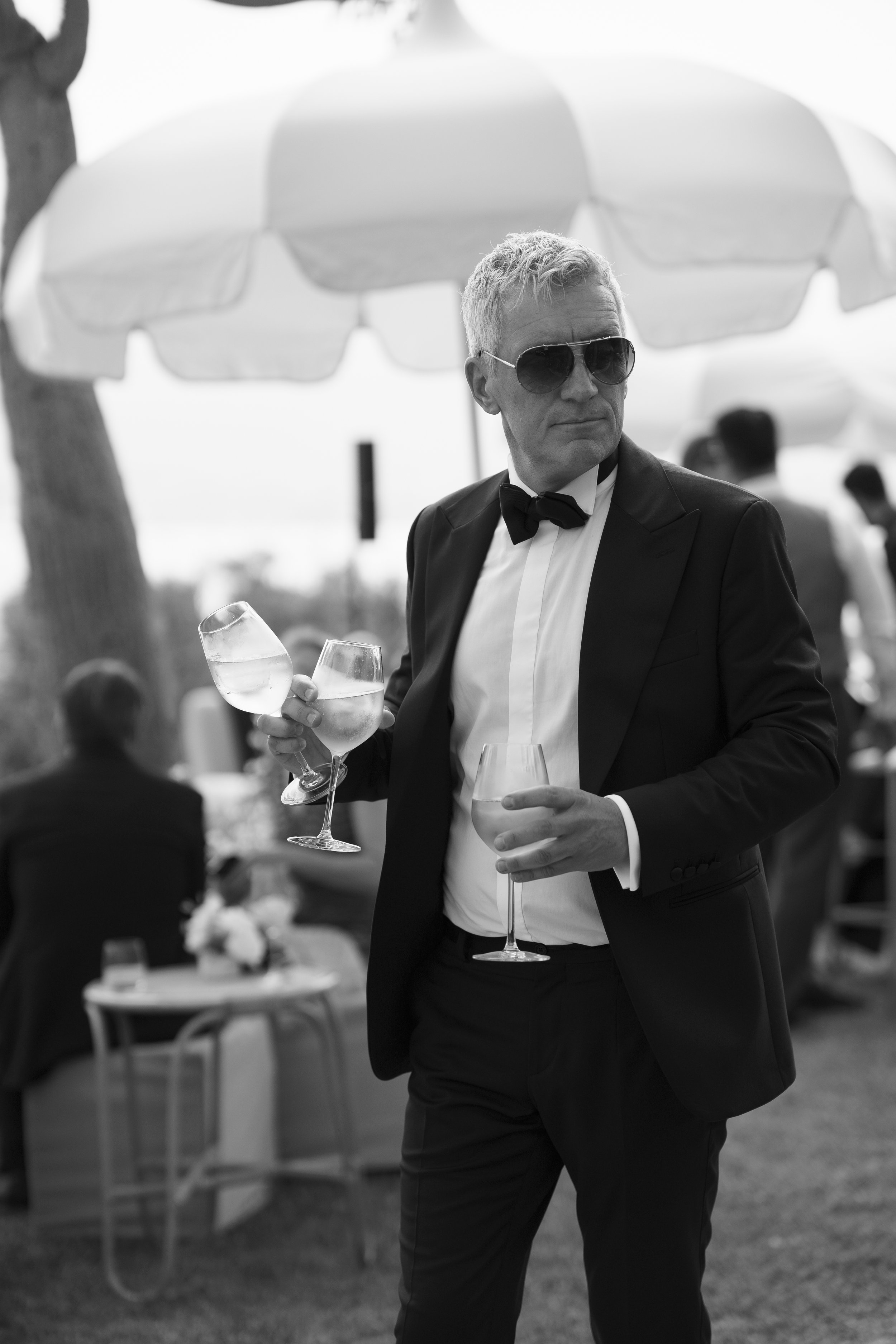 A man in a tuxedo with sunglasses holding three glasses of wine at an outdoor event with umbrellas.