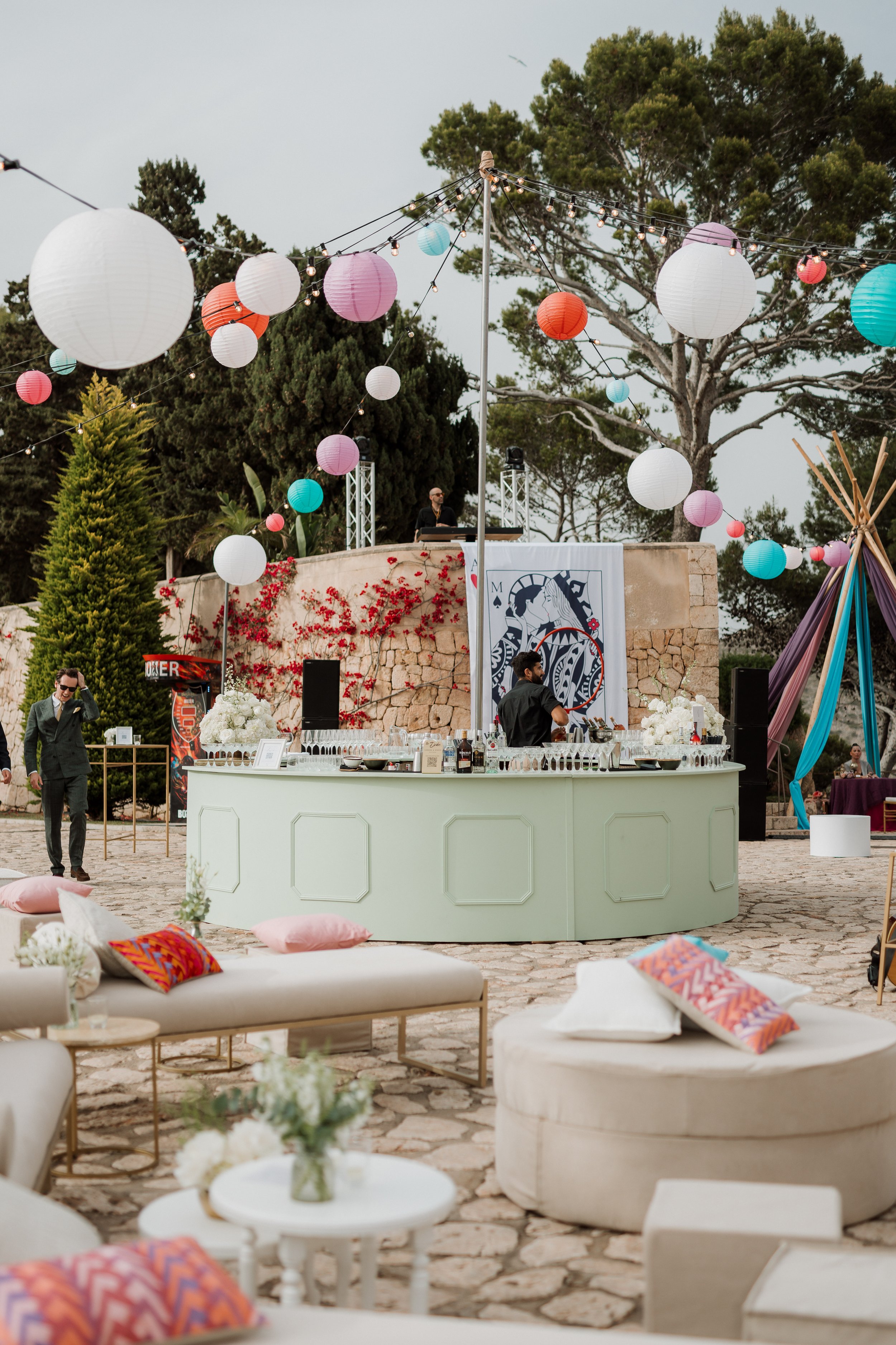 An outdoor party setup with hanging paper lanterns and string lights, a bar with drinks, a DJ booth on a raised platform, and cushioned seating with decorative pillows, surrounded by trees and a stone wall.