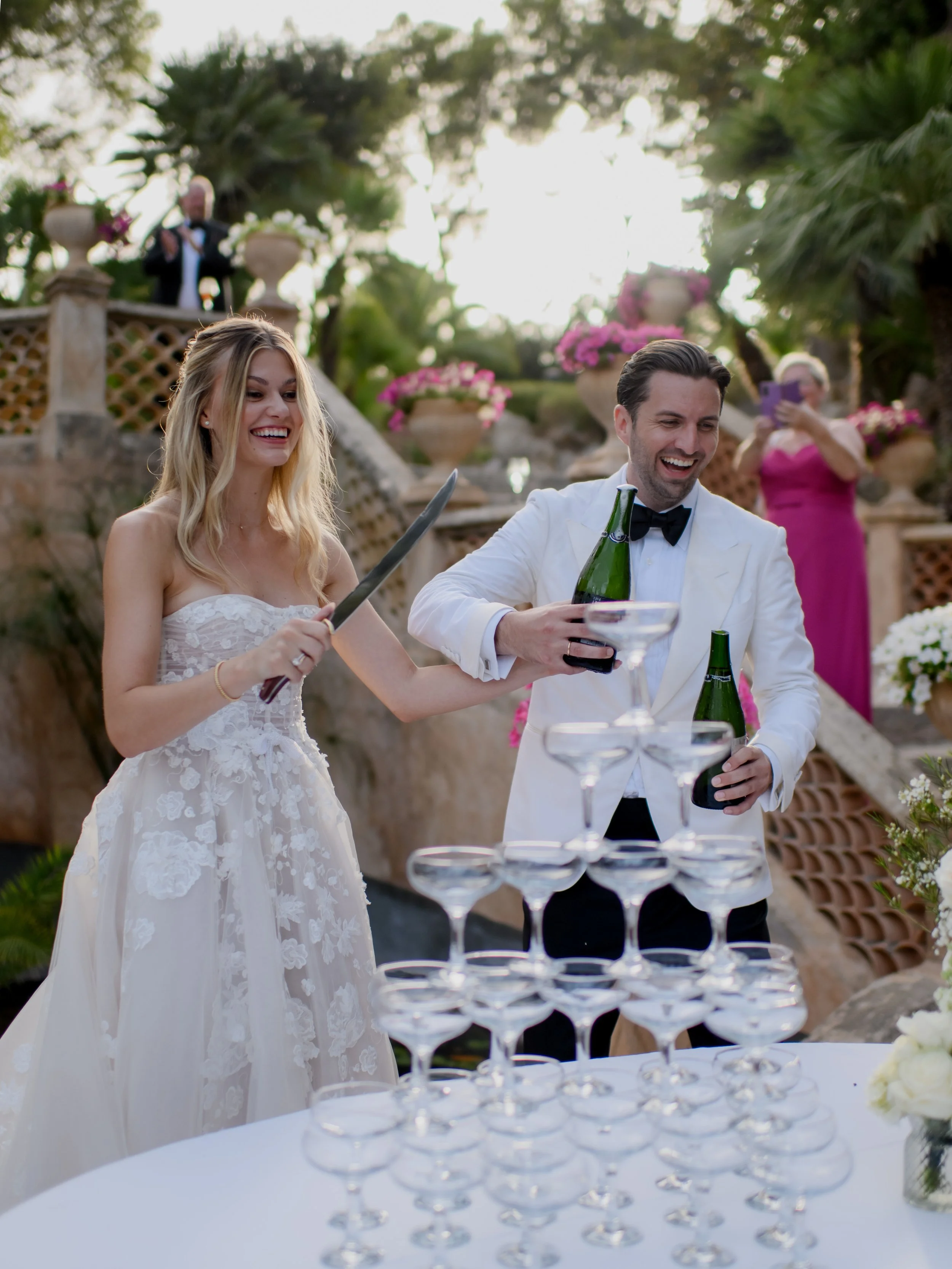 A bride and groom are standing outdoors during their wedding celebration, smiling and pouring champagne into a tower of glasses, with friends and guests taking photos in the background.