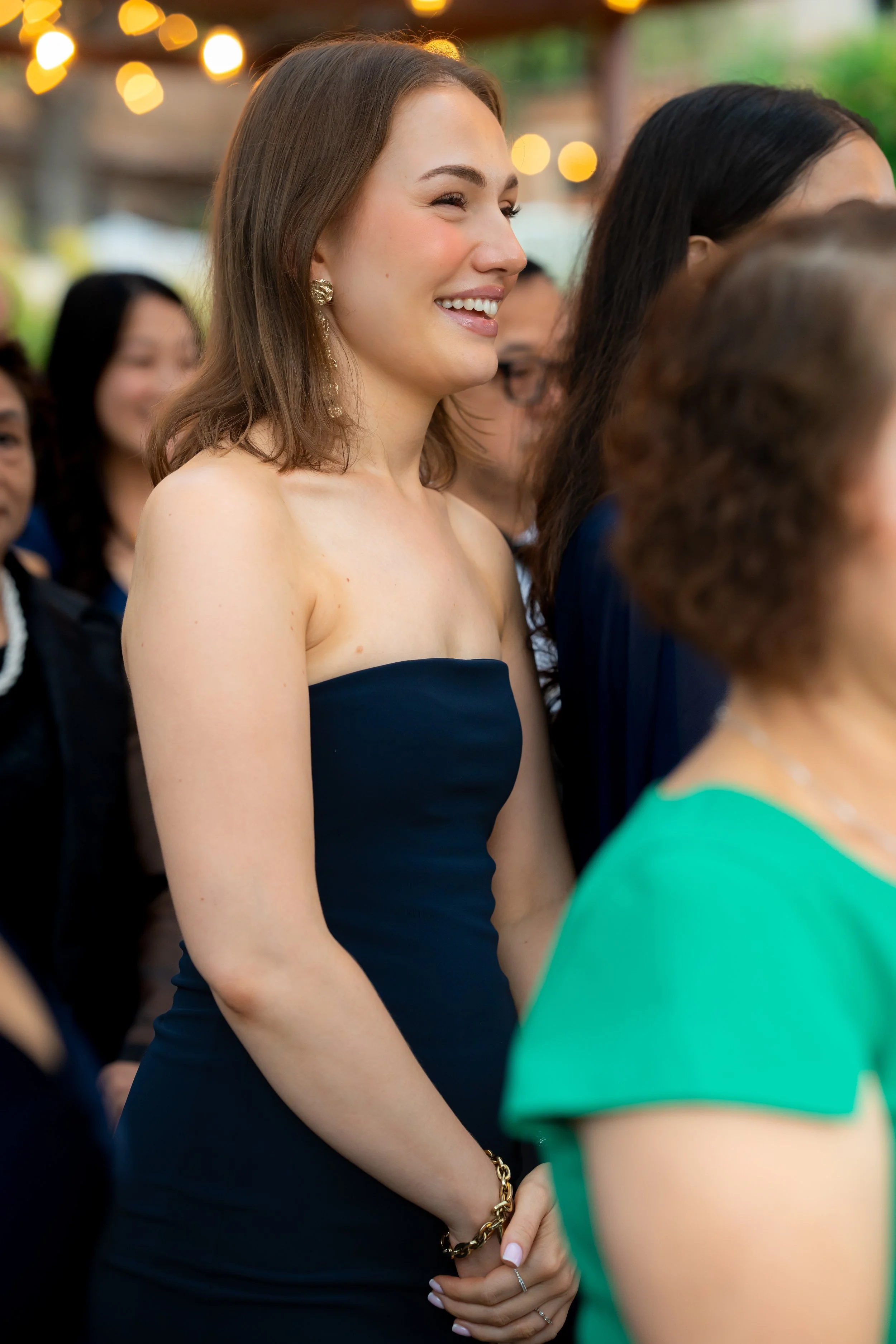 A woman with shoulder-length brown hair wearing a strapless dark blue dress, gold earrings, a gold bracelet, and a ring, smiling at an outdoor event with string lights in the background.