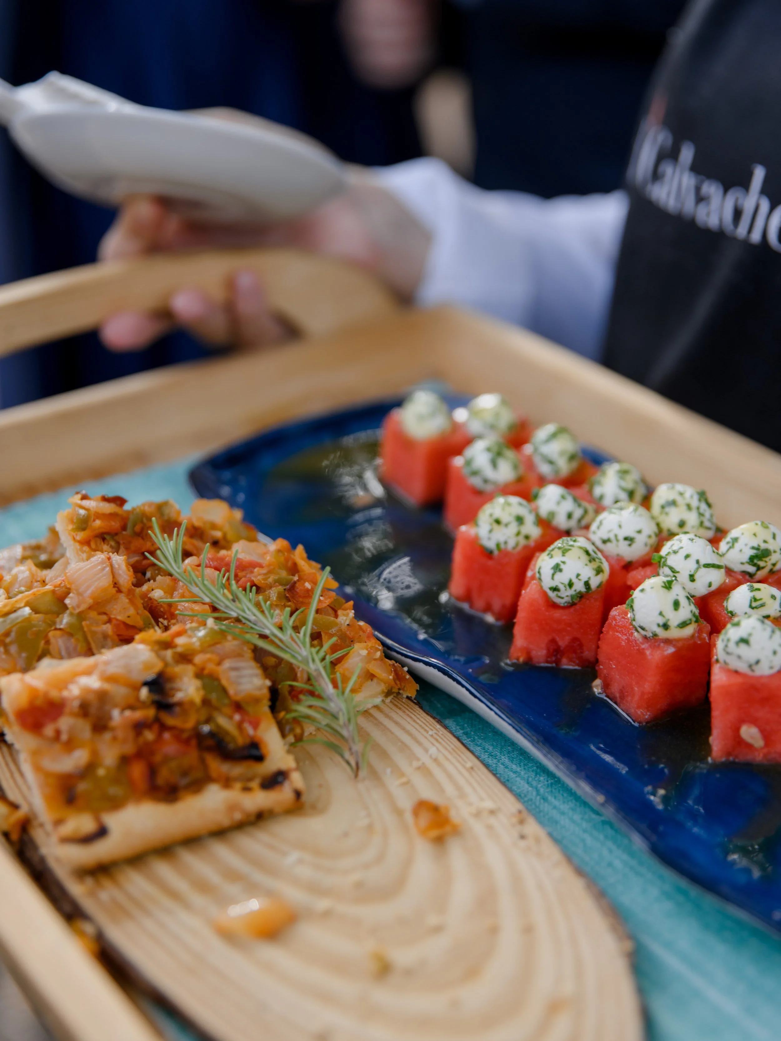 Watermelon cubes topped with dollops of herb-infused cream. A slice of vegetable quiche garnished with a sprig of rosemary, served on a wooden tray.
