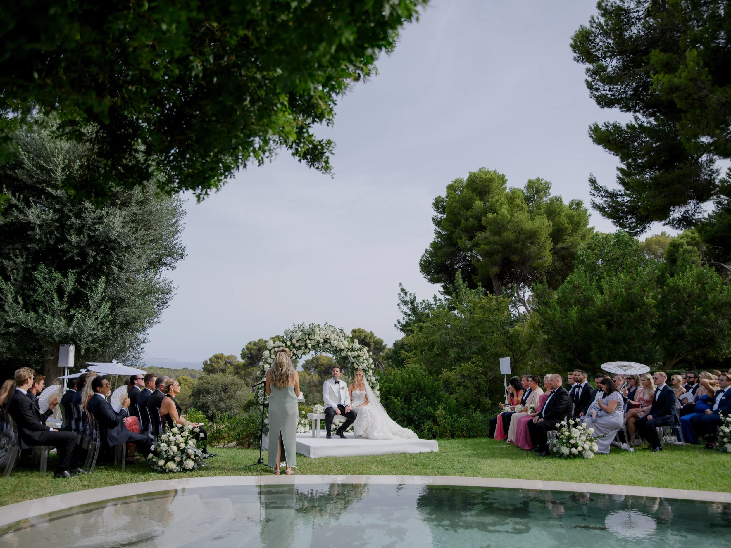 Outdoor wedding ceremony with bride and groom seated at altar decorated with white flowers, officiant standing, surrounded by seated guests, green trees, and a reflective pool in the foreground.