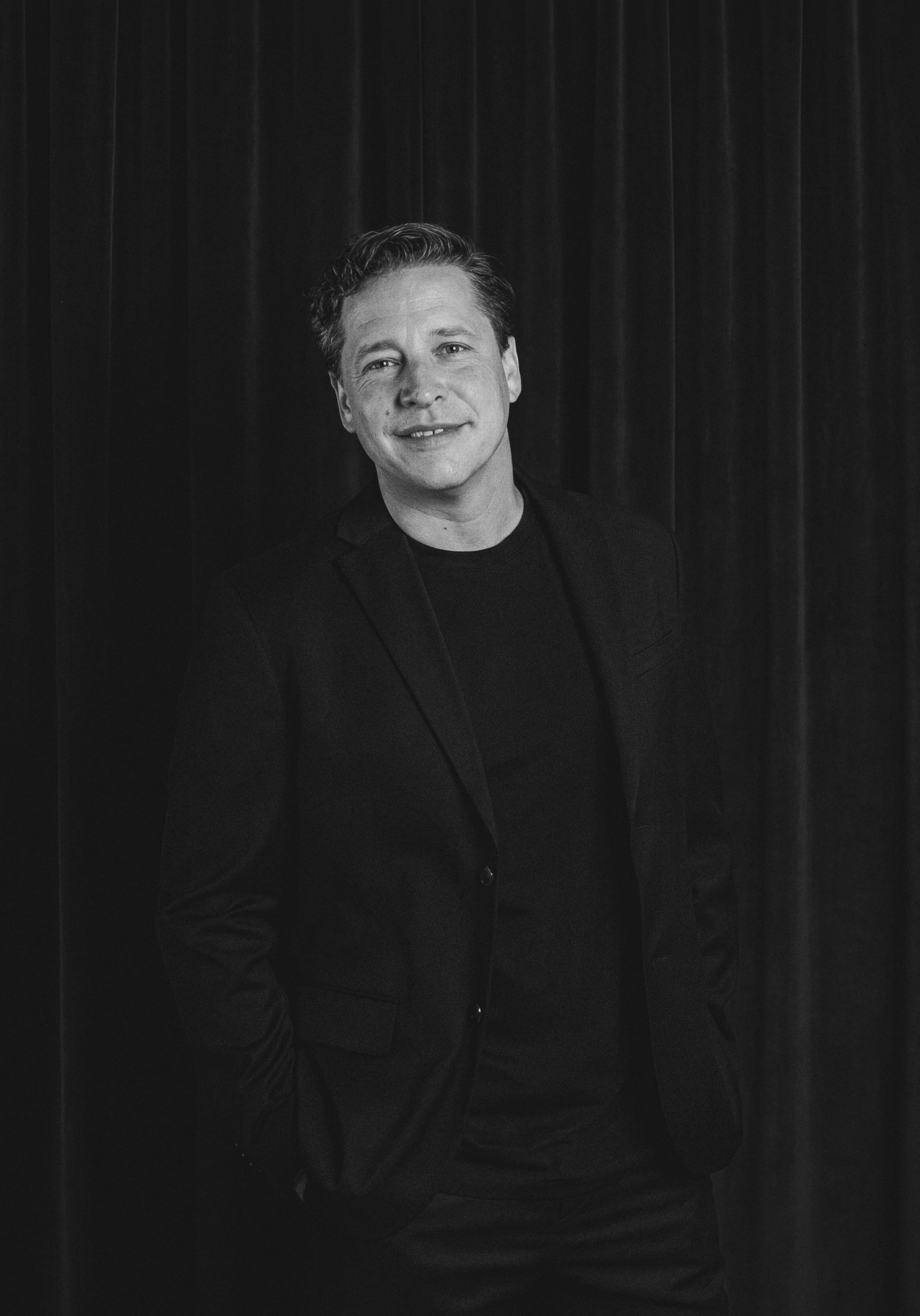 A man with curly hair in a black blazer standing against a dark curtain background, smiling at the camera.