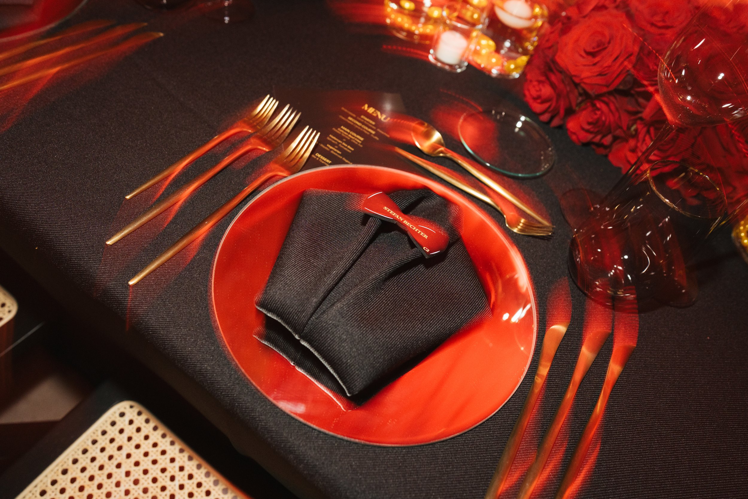 Elegant tablescape with red and black color scheme featuring a red plate with a folded black napkin, gold utensils, red roses, wine glasses, candles, and a menu on a black tablecloth.