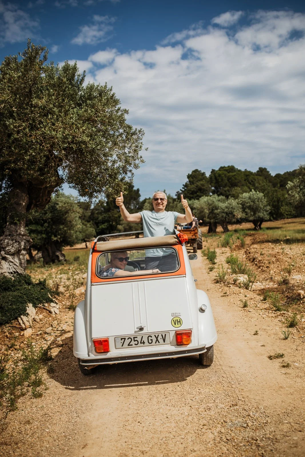 Two people in a vintage white and orange microcar on a dirt path surrounded by olive trees, with one person standing through the roof giving a thumbs-up and the other sitting inside smiling.