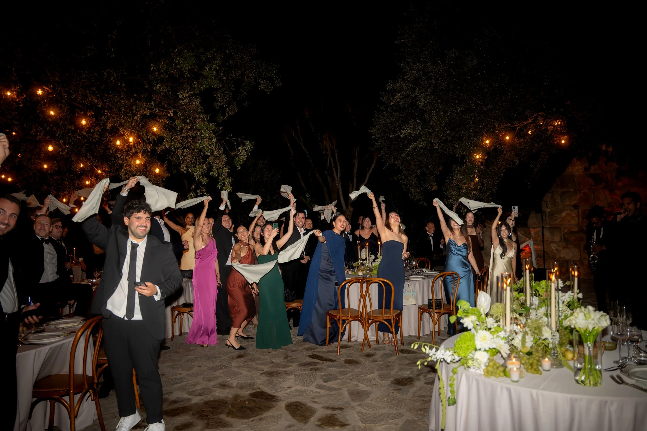 Guests at a wedding reception dancing and celebrating outdoors at night with decorated tables and string lights.