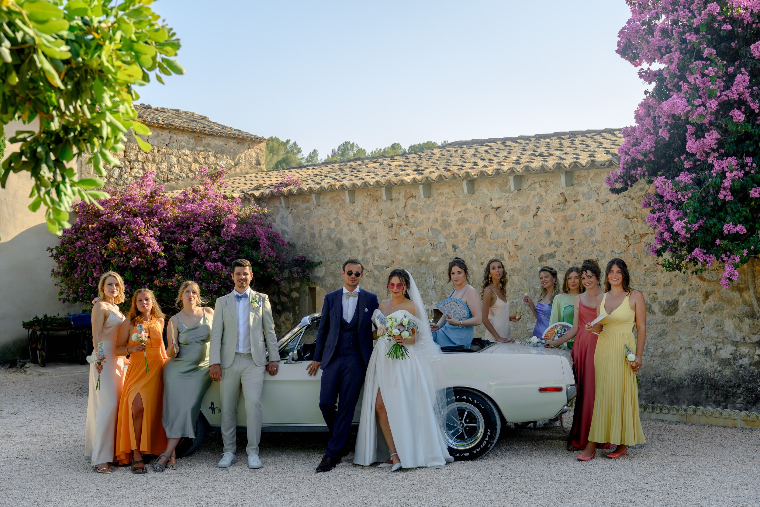 A wedding party with the bride and groom in front of a vintage white convertible car, surrounded by bridesmaids and friends, in a rustic outdoor setting with stone walls, flowering bushes, and clear weather.