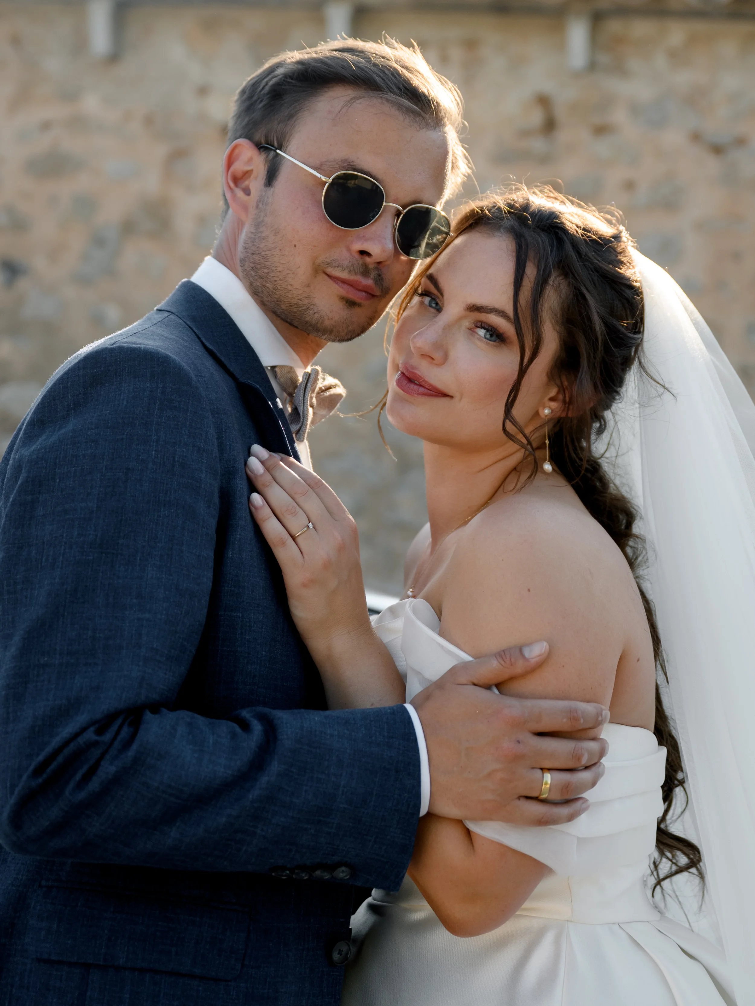 A bride and groom in wedding attire standing close together, with the bride wearing an off-the-shoulder wedding dress and veil, and the groom in a navy suit and sunglasses, against a stone wall background.