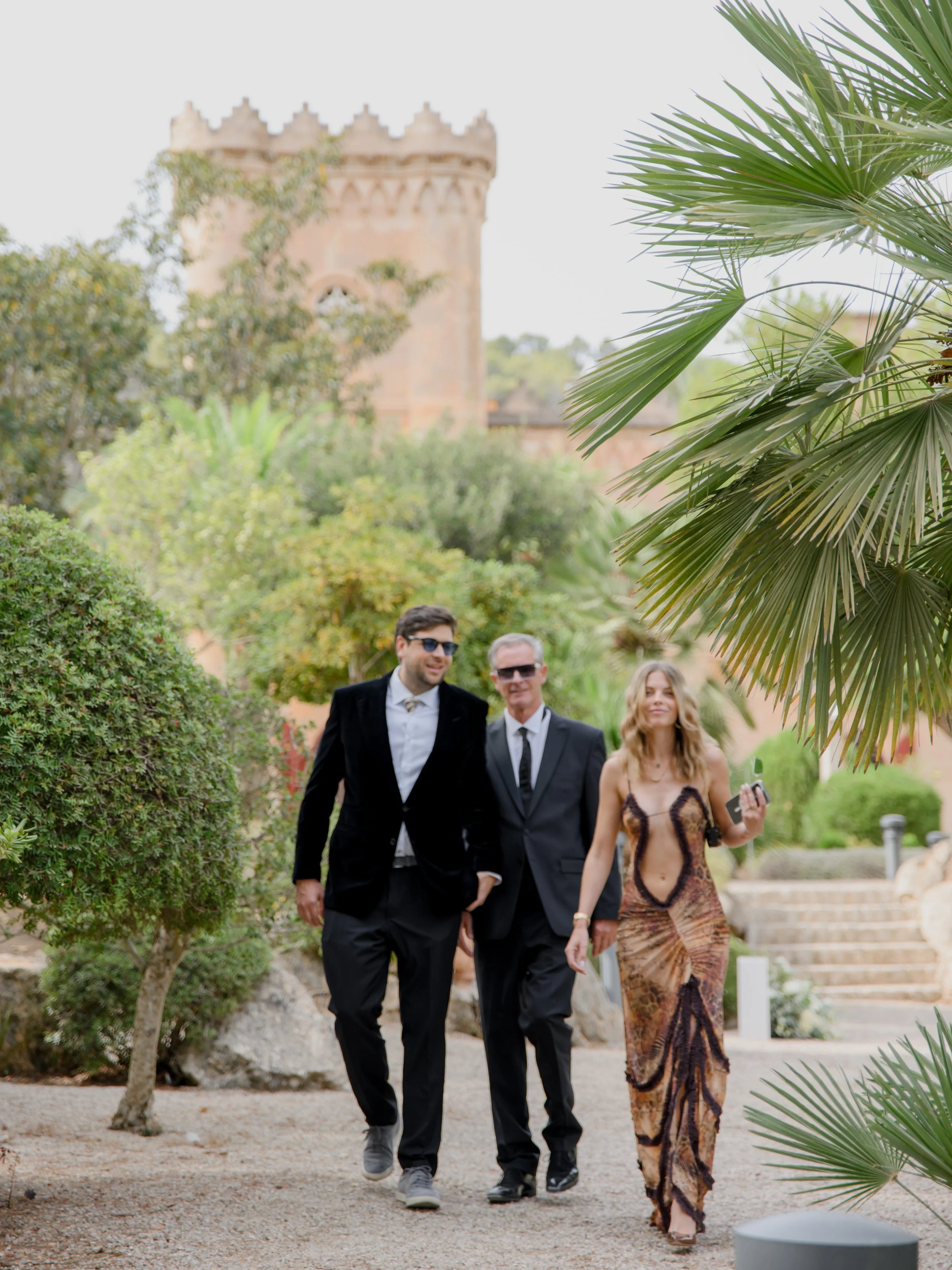 Three people walking outdoors on a garden pathway, with lush green trees and a castle-like tower in the background. Two men are in black suits, and a woman is in a colorful, patterned dress, holding a phone.