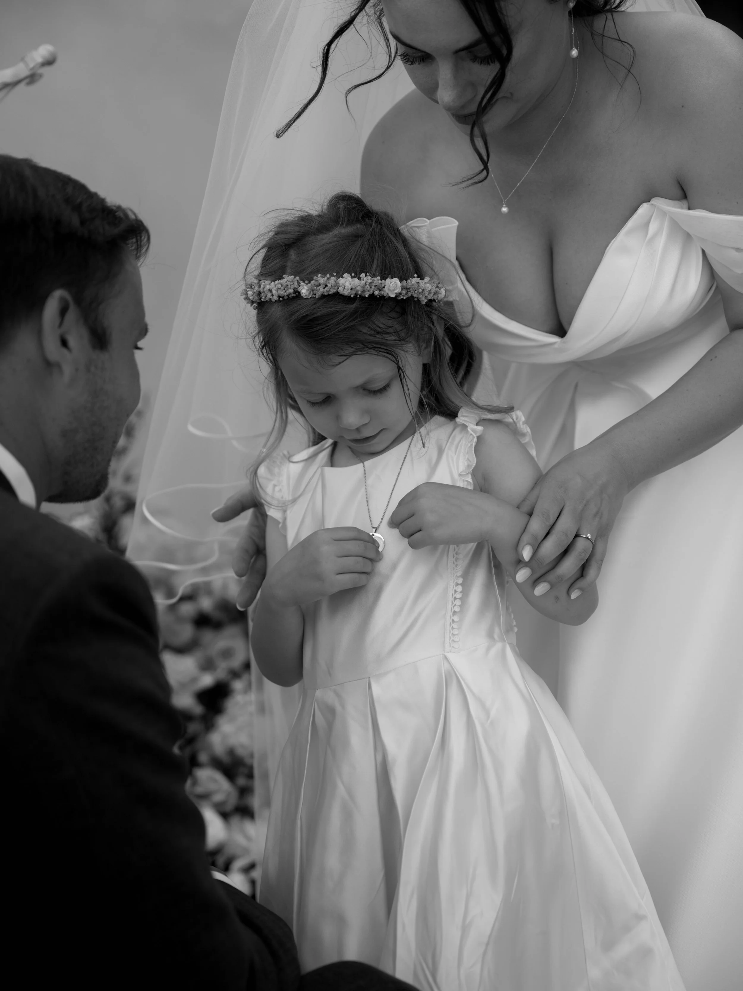 A woman in a wedding dress and a little girl in a dress are holding jewelry, with a man kneeling before them during a wedding ceremony.