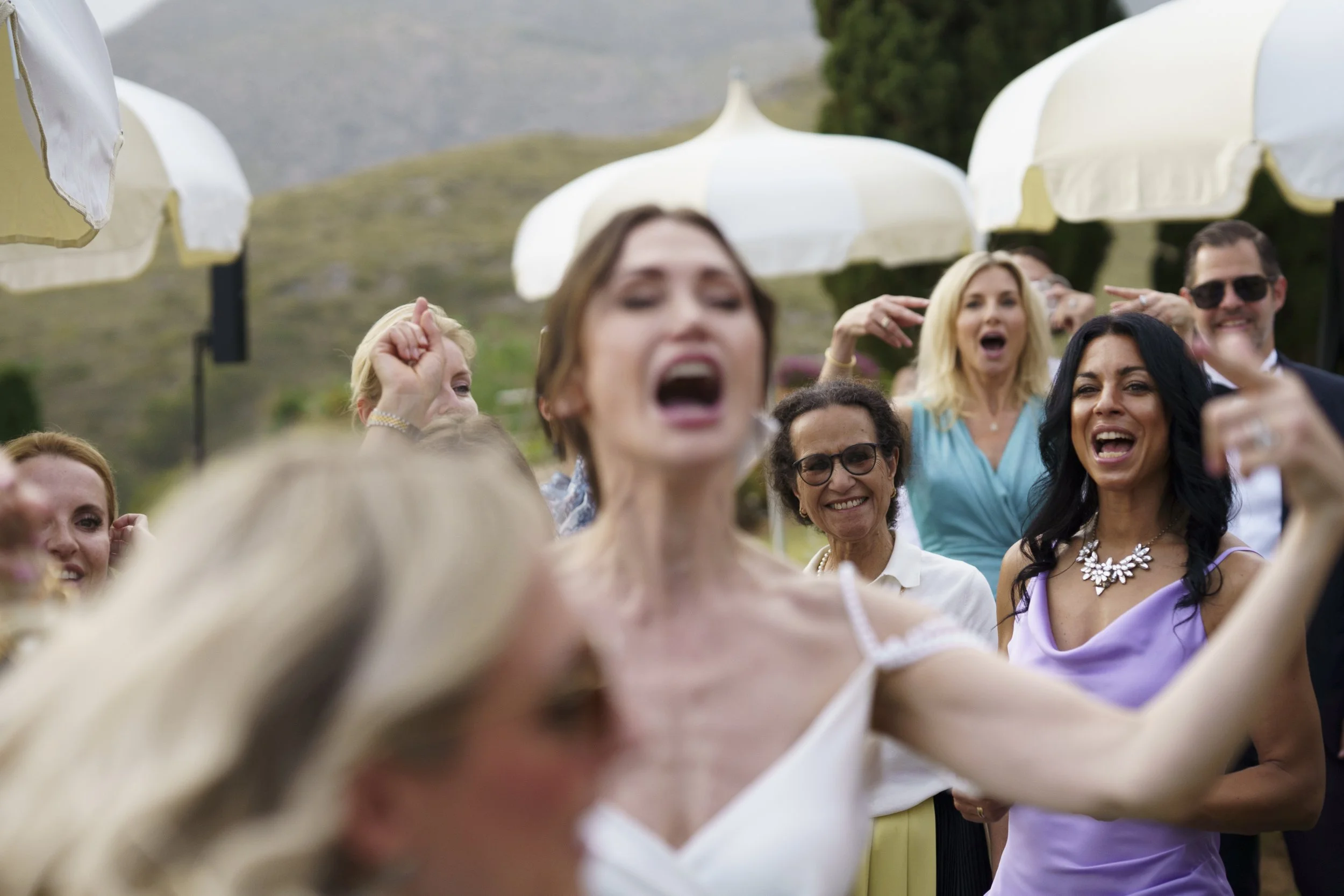 A group of women and one man at an outdoor event, smiling and raising their hands, with white umbrellas and a hilly background.