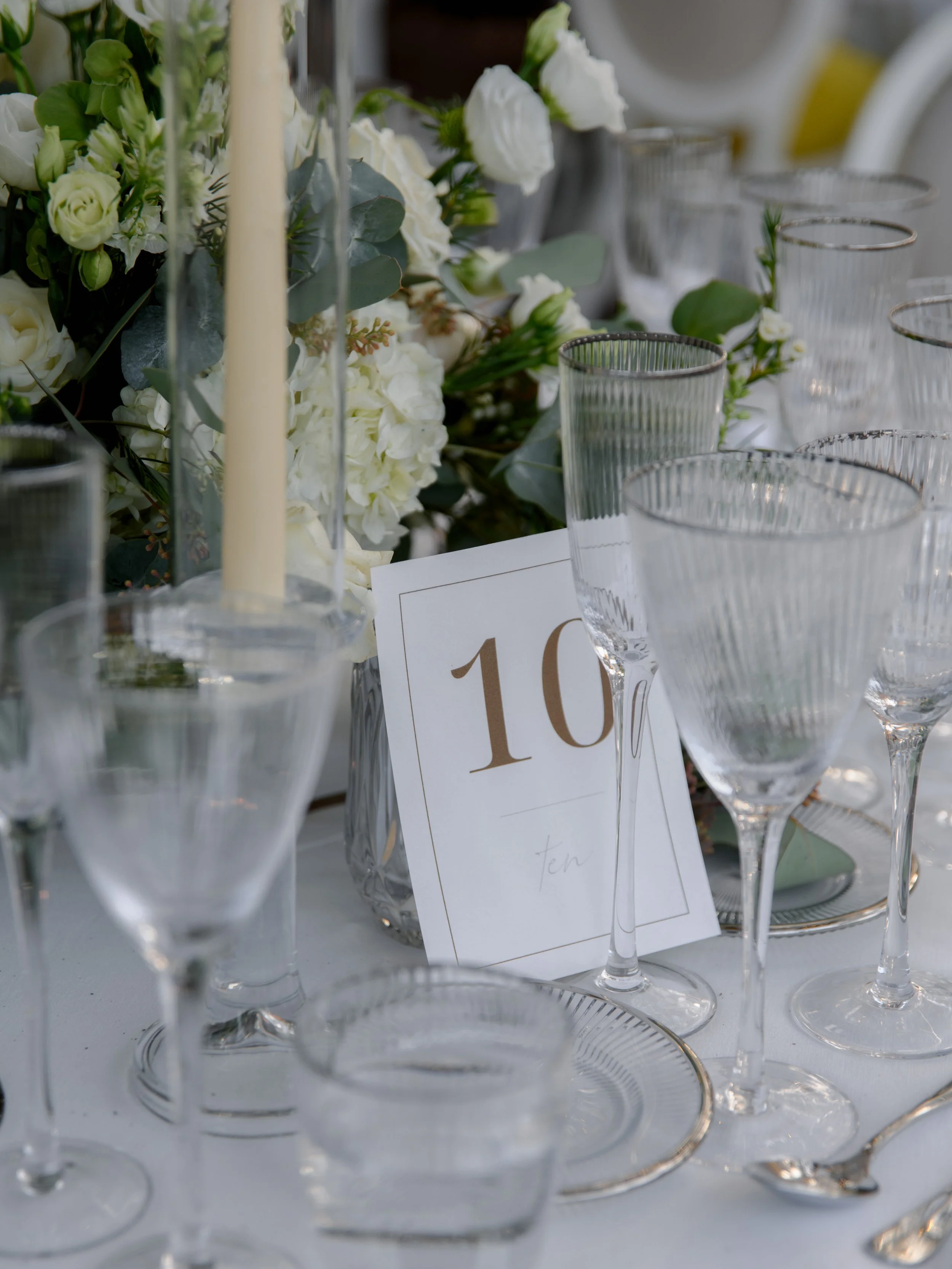 Elegant table setting with multiple clear glassware including wine glasses, a floral centerpiece with white flowers and greenery, a white tablecloth, and a table number card displaying the number 10.