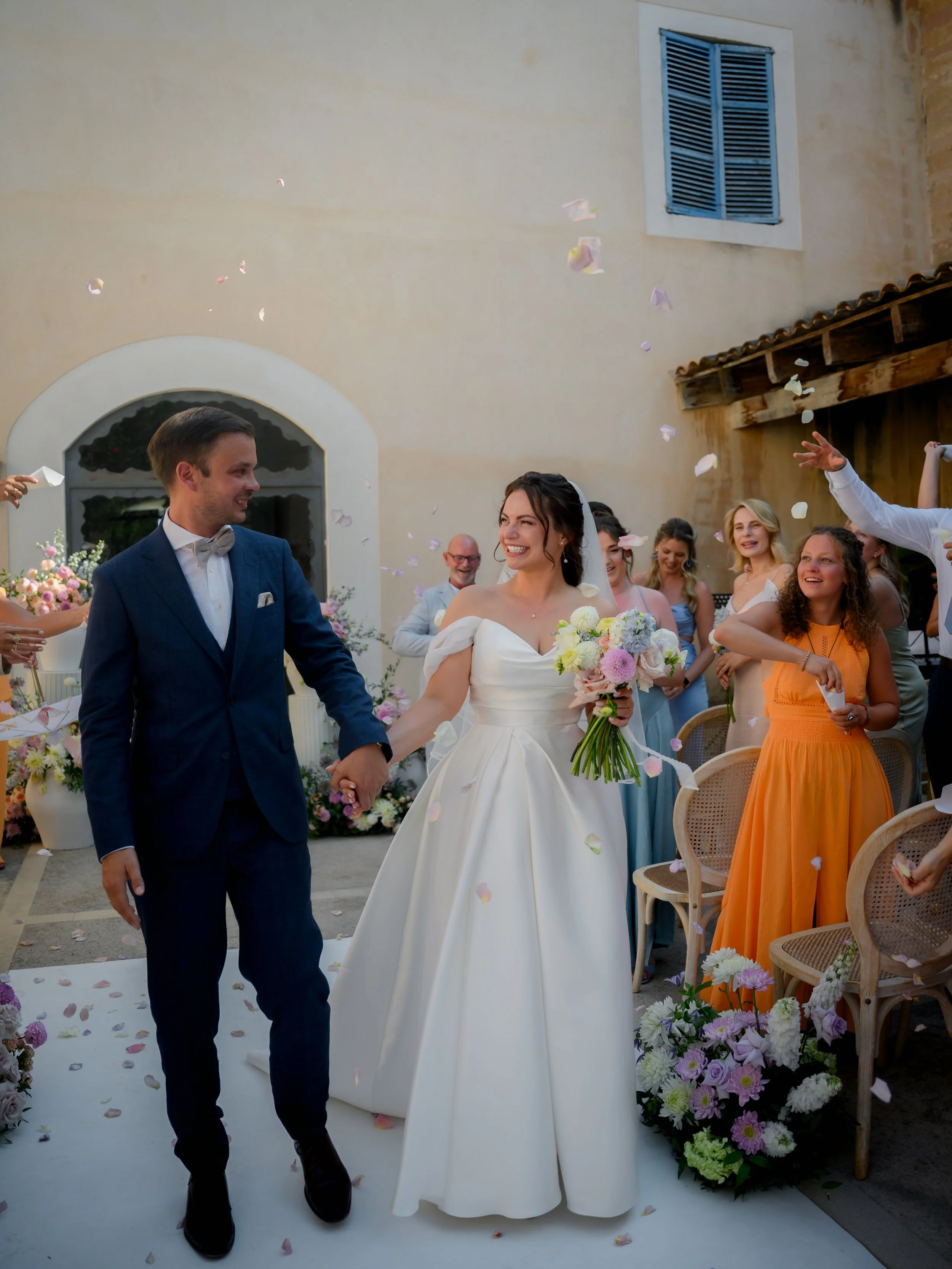 Bride and groom holding hands while walking down the aisle at their outdoor wedding surrounded by friends and family, with flower petals in the air.