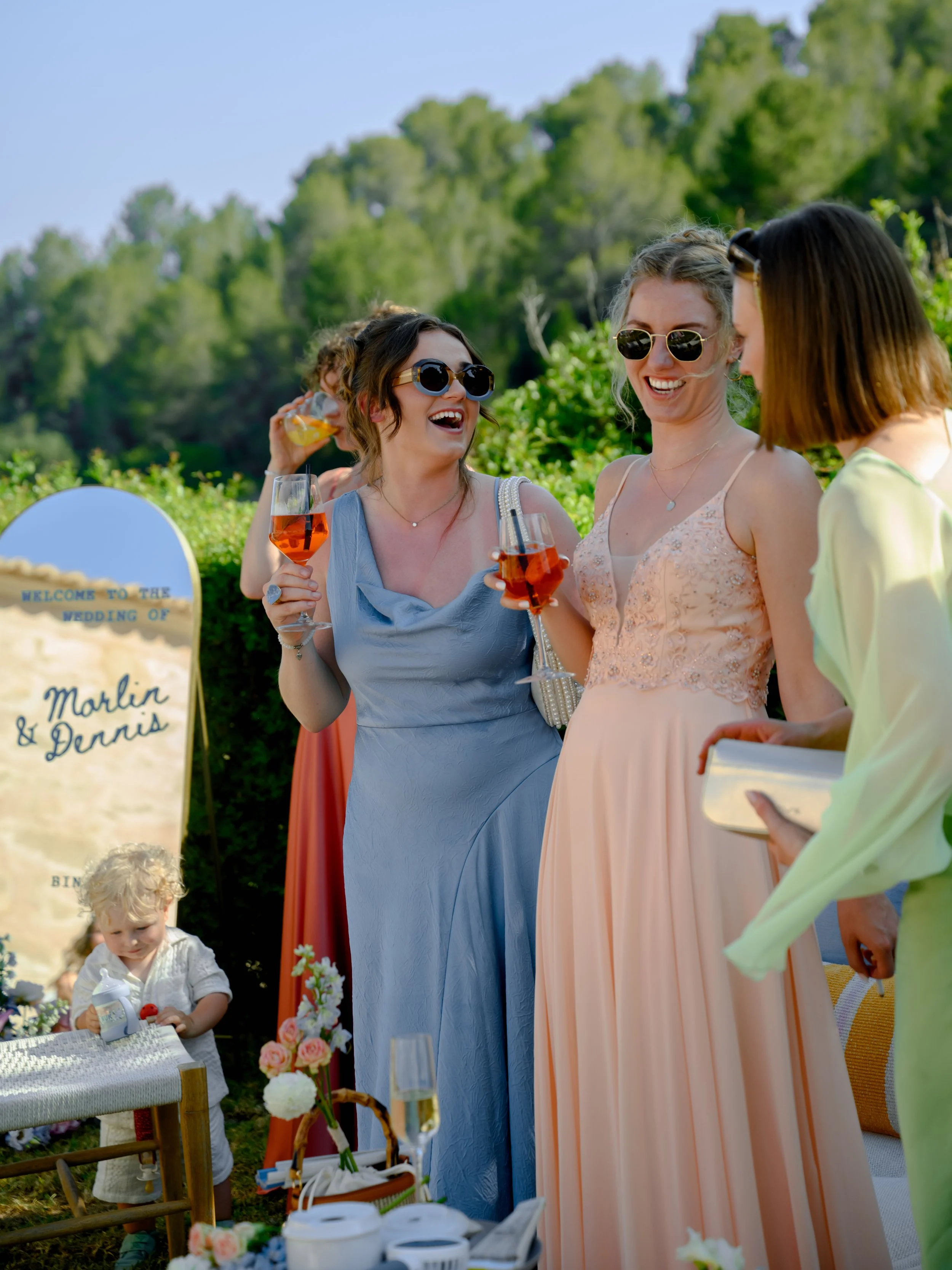 Three women in colorful dresses and sunglasses celebrating with drinks outdoors at a wedding reception, with a young child and floral decorations in the background.