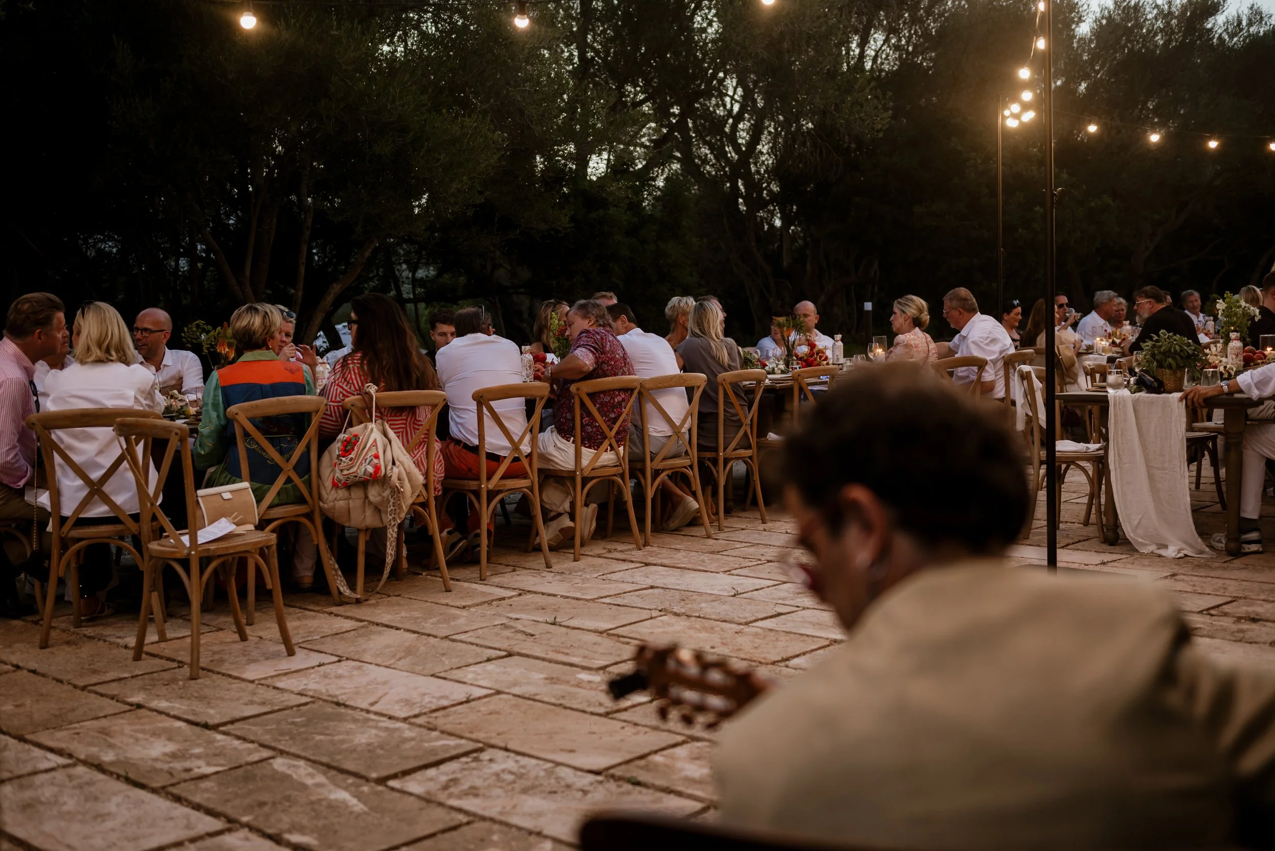 People gather around long dinner tables under string lights outdoors during evening