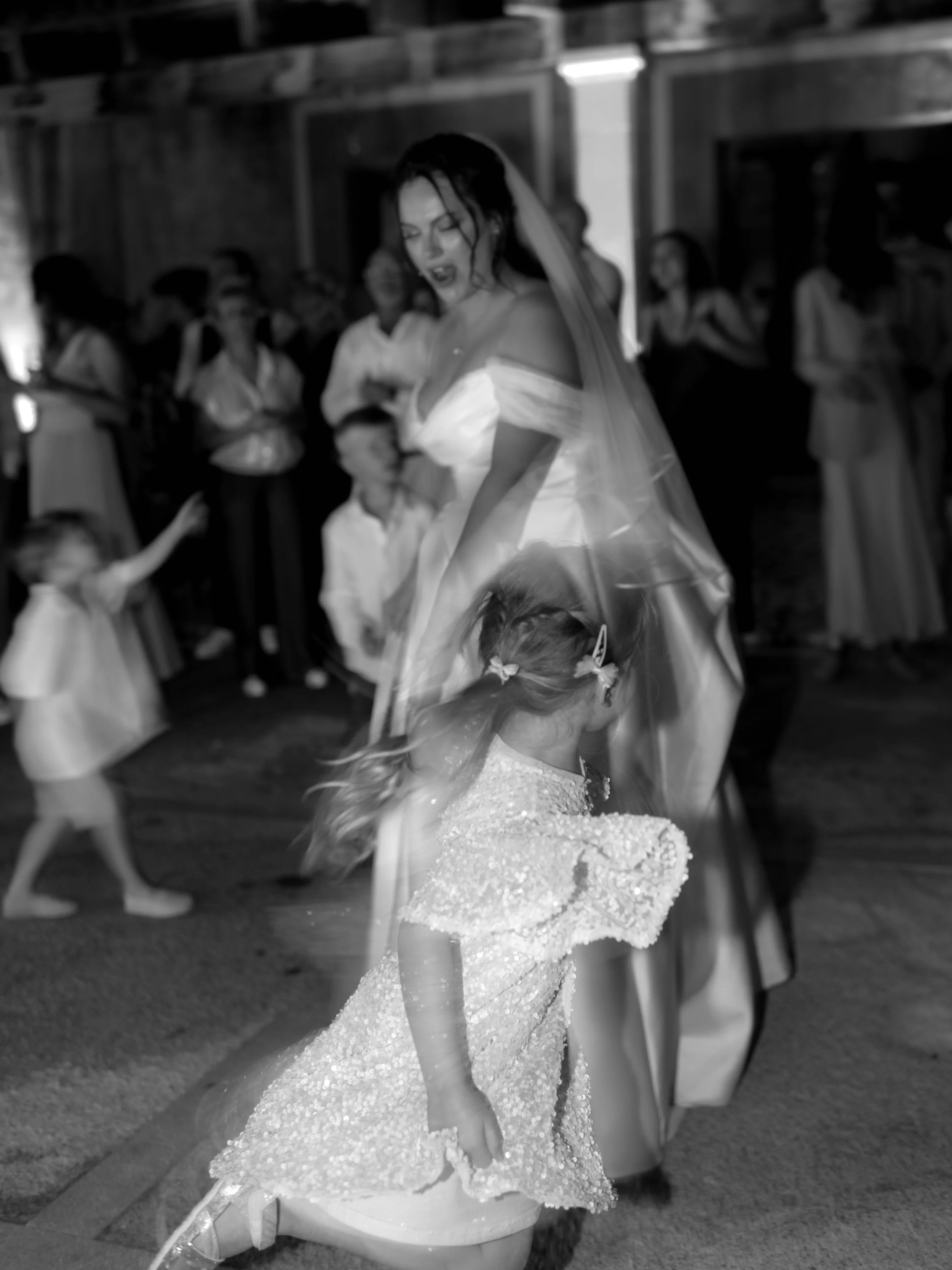 Black and white photo of a bride dancing with a young girl, surrounded by guests at a celebration or wedding reception.