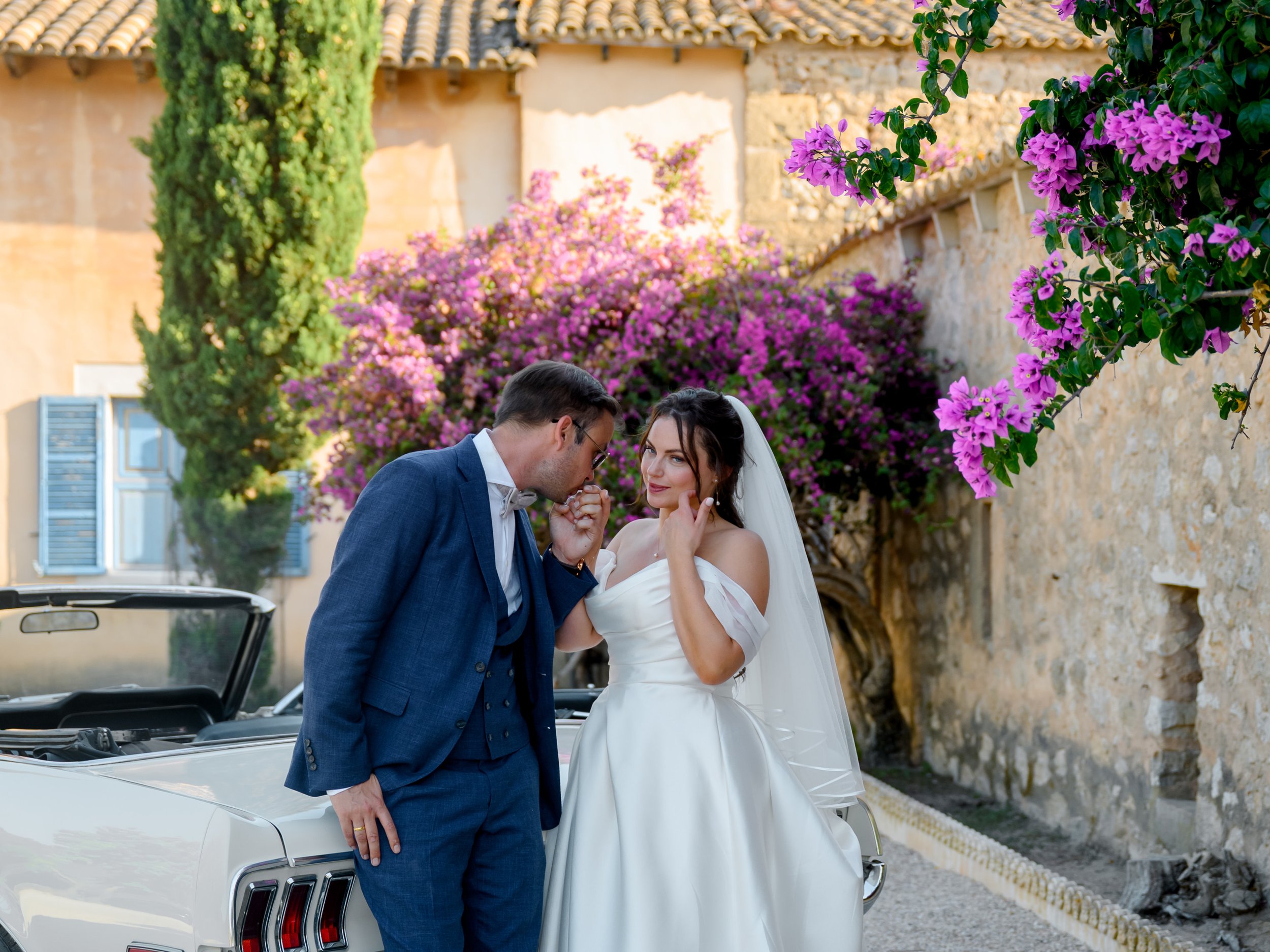 A bride and groom standing near a vintage white convertible car, outdoors with colorful flowers and an old stone building in the background. The groom is dressed in a blue suit and sunglasses, leaning towards the bride, who is wearing a white wedding