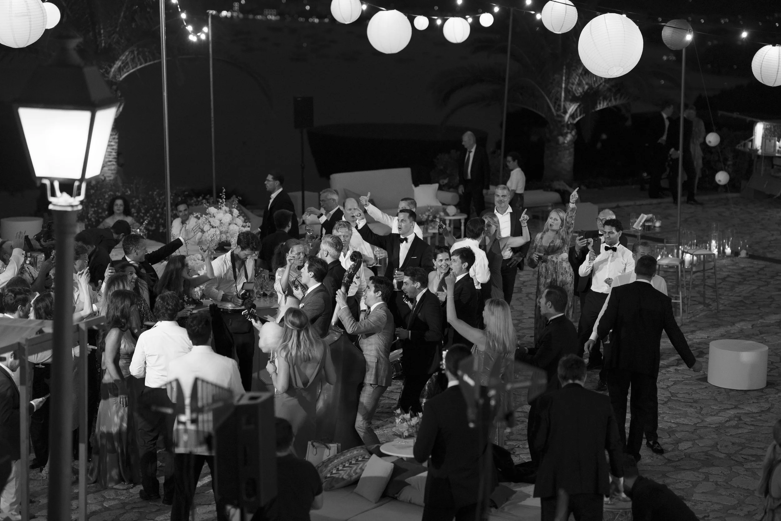 A black and white photo of a lively outdoor party with many people dancing and socializing under hanging paper lanterns at night.