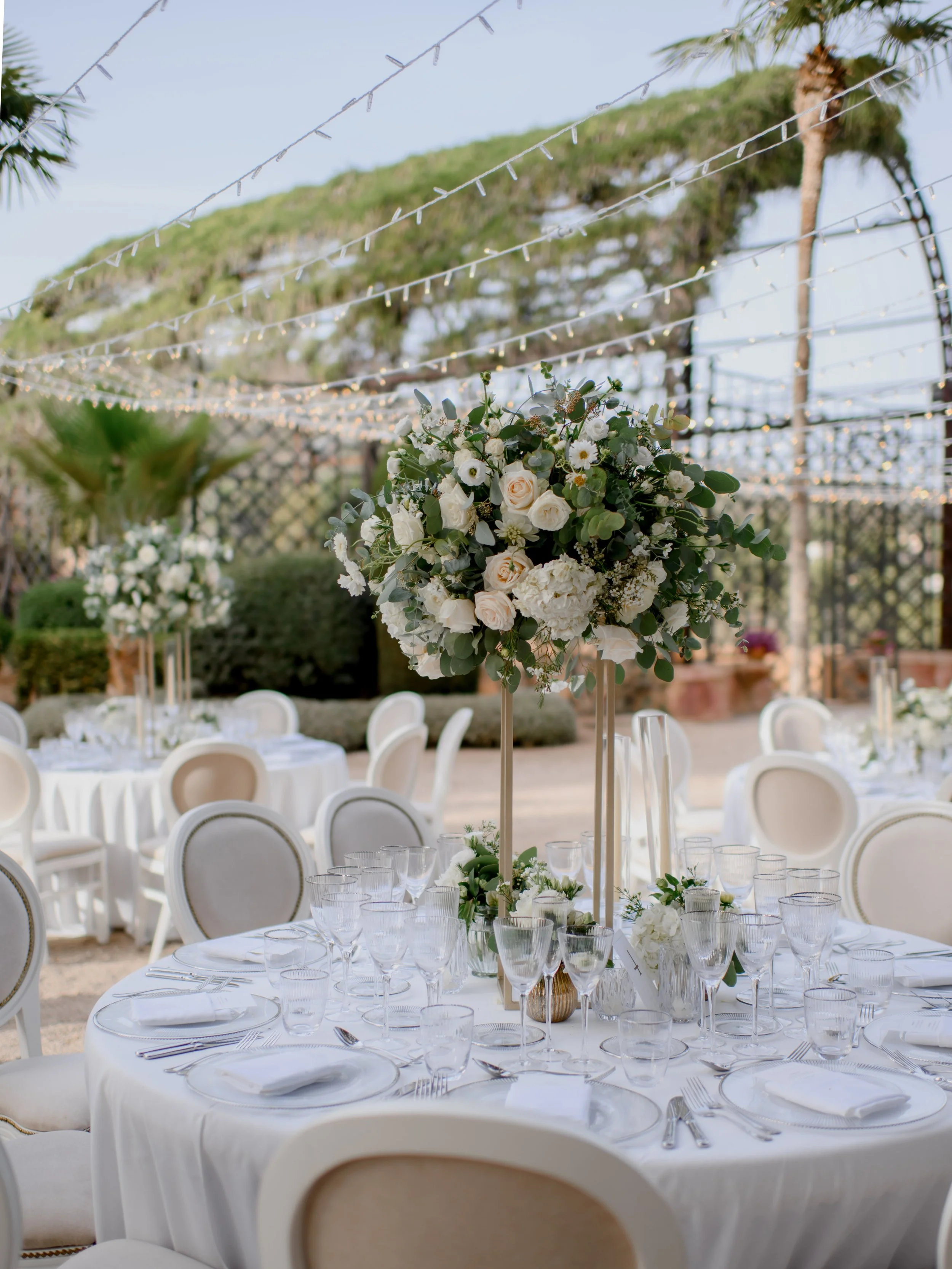 Elegant outdoor event setup with round tables, white tablecloths, and floral centerpieces, under string lights with palm trees and greenery in the background.