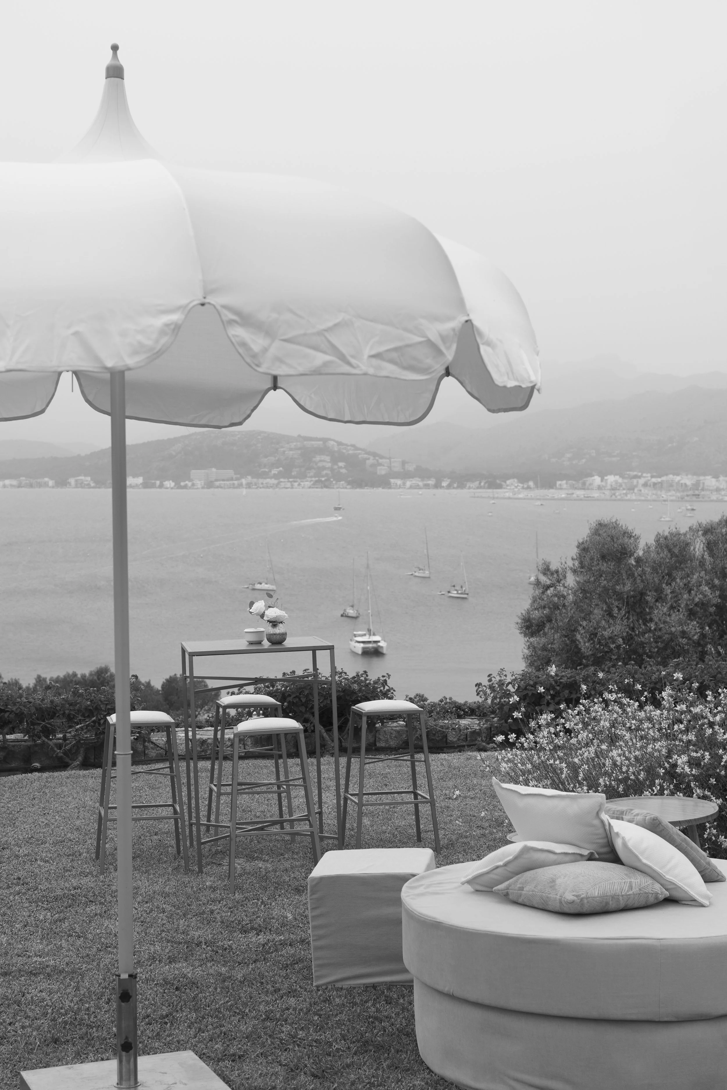 Outdoor patio area with a large umbrella, a round cushioned seating area with pillows, a small table, and a high table with bar stools, overlooking a scenic water view with sailboats and distant hills.