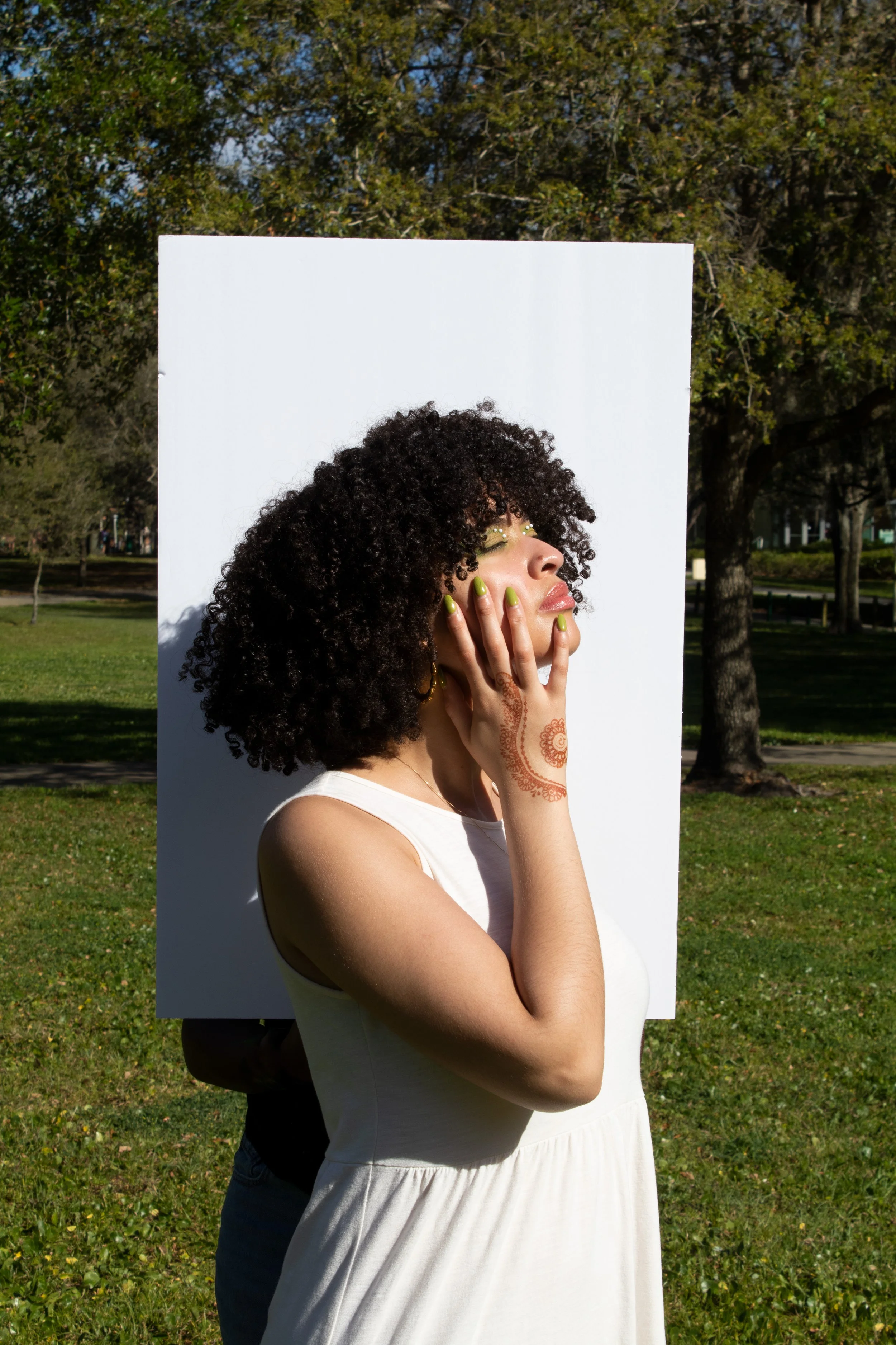 A woman with curly hair, wearing a white sleeveless dress, standing outdoors in a park with trees in the background. She has henna on her hand, green nail polish, and gold makeup, and is touching her face with her hand.