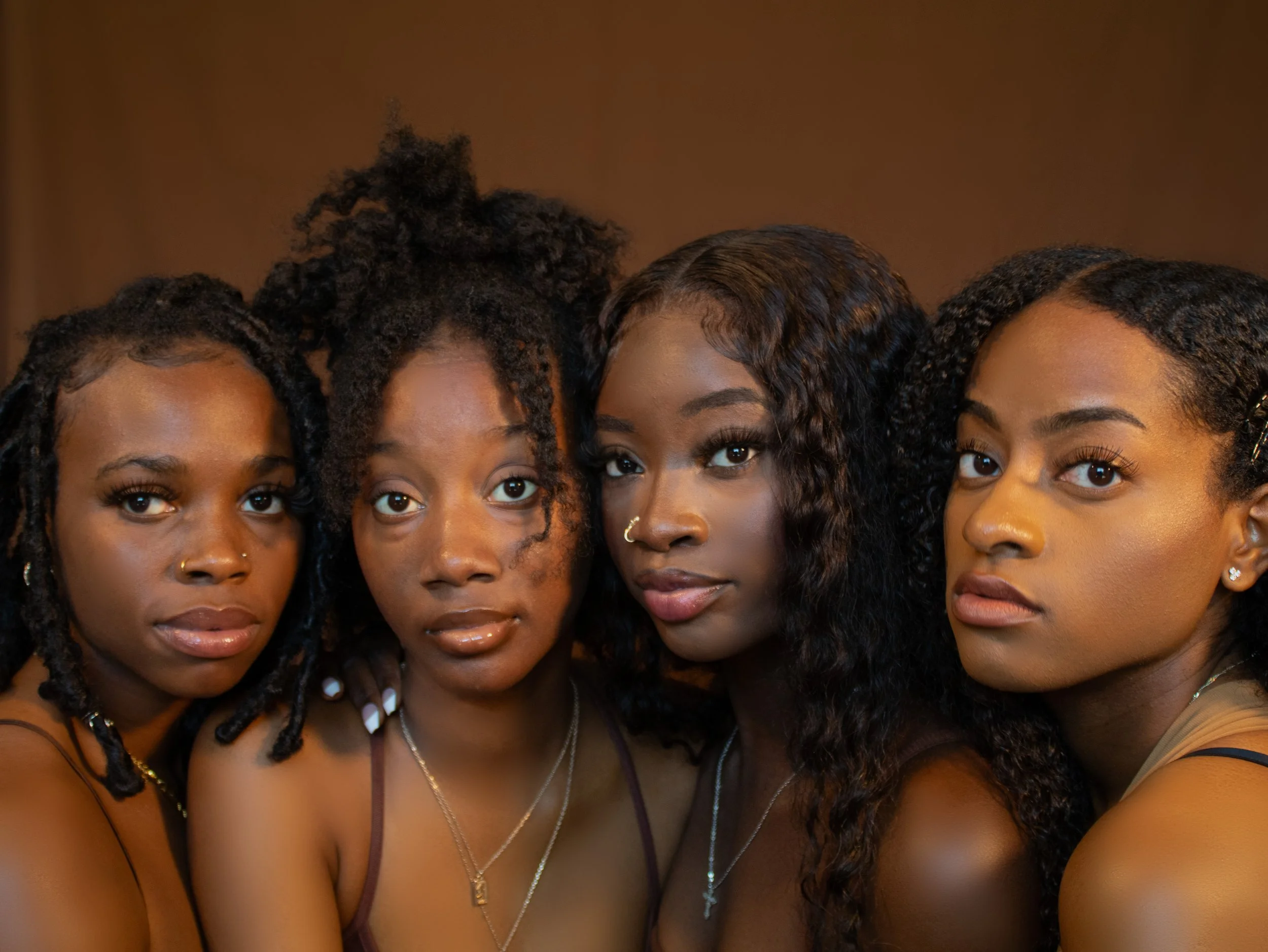 Four women posing closely for a photo against a brown background, with diverse hairstyles and wearing necklaces and earrings.
