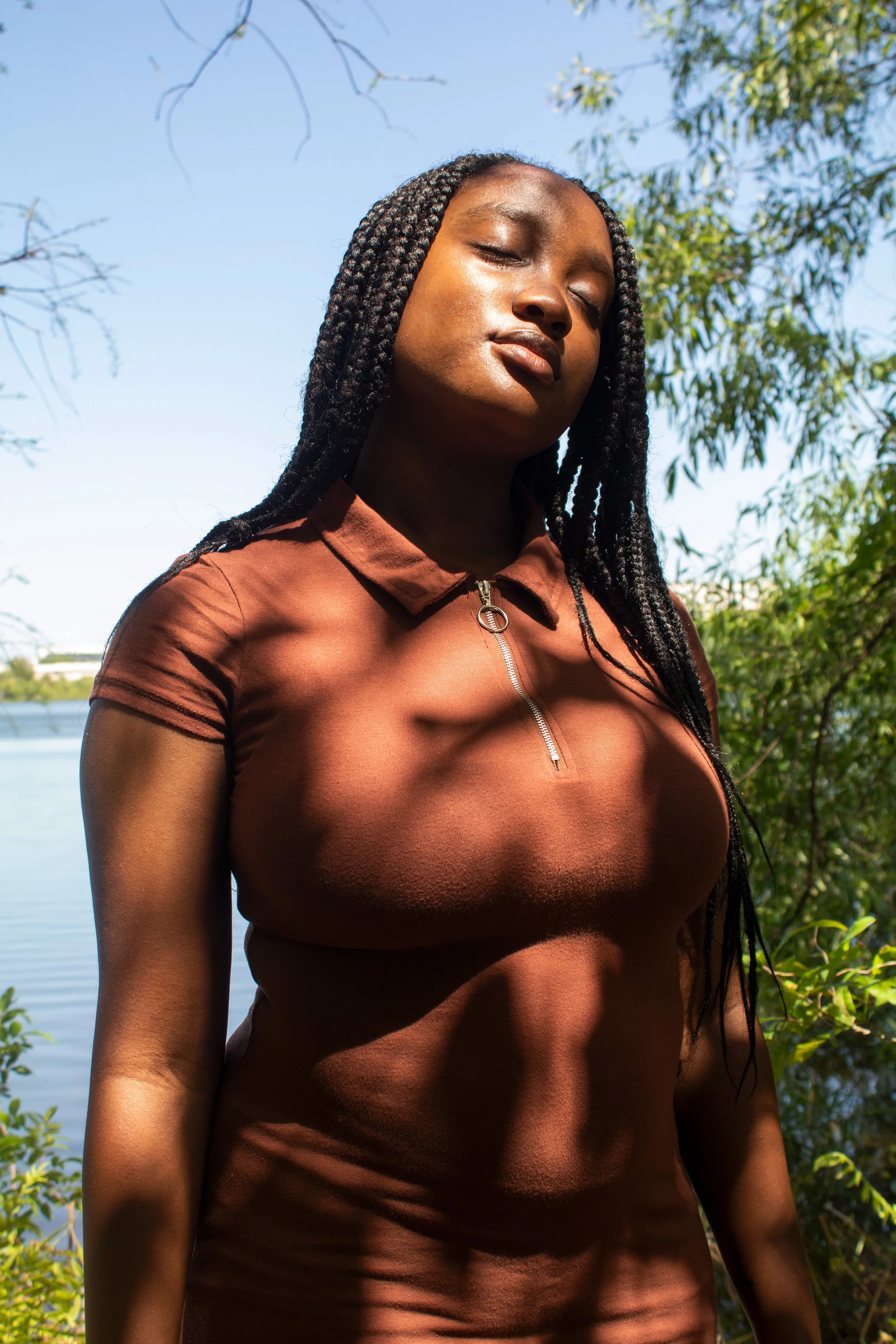 A woman with braided hair standing outdoors beside a body of water with trees and blue sky in the background.