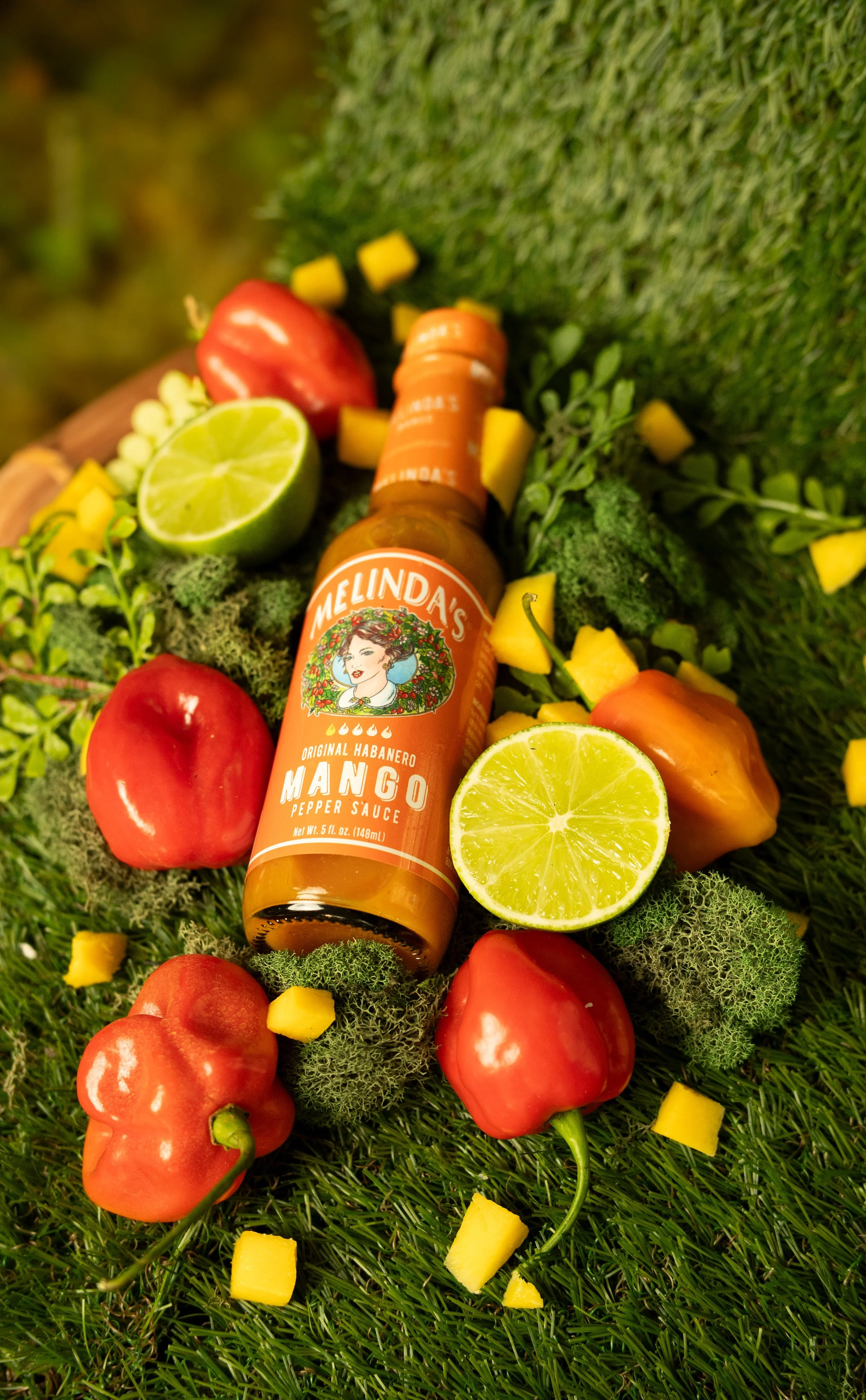 A bottle of Melinda's Mango hot sauce surrounded by fresh vegetables and fruits, including red bell peppers, jalapeño peppers, lemon halves, and small yellow diced pieces, all arranged on a bed of green grass and moss with small leaves.