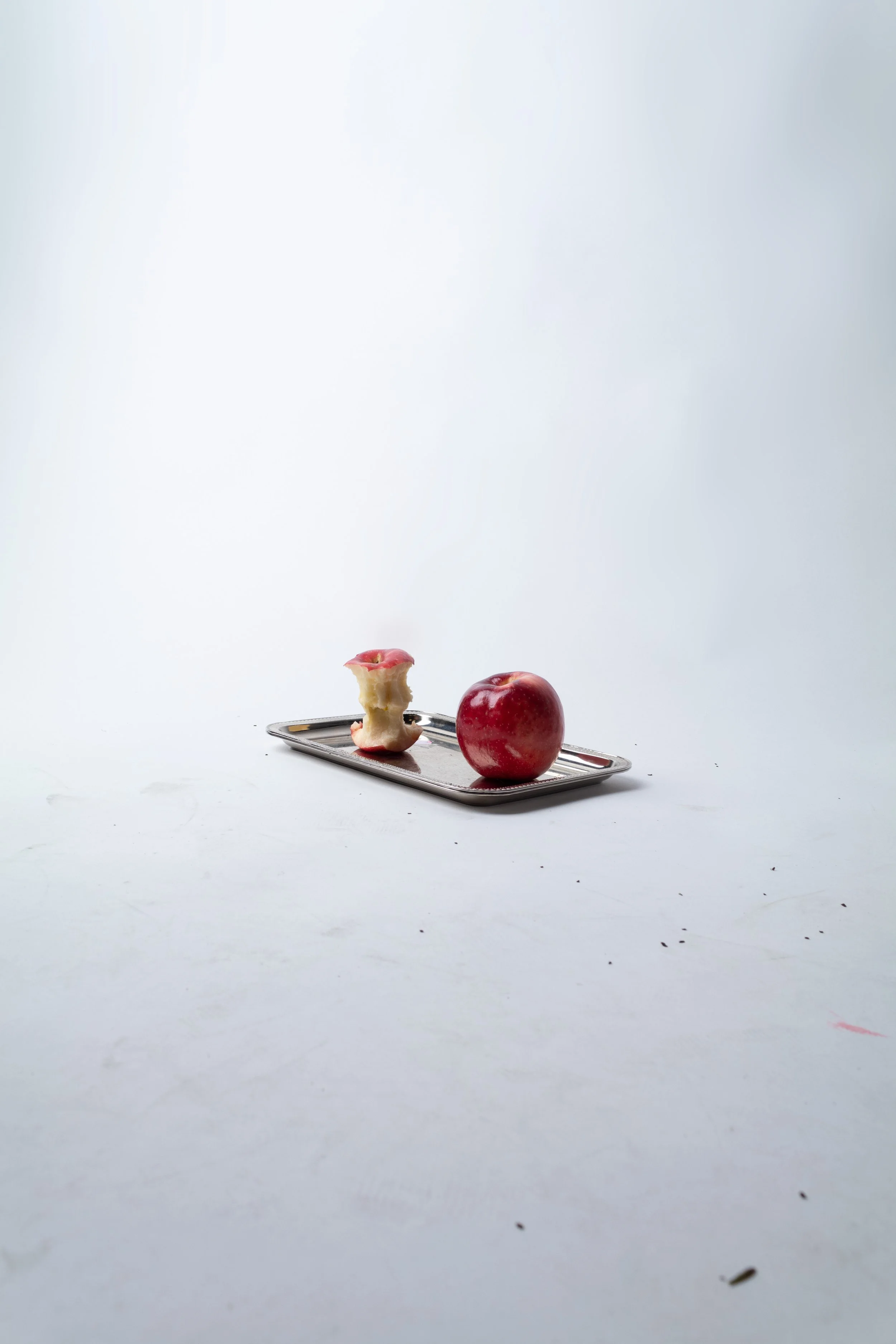 A partially eaten red apple with a bite taken out of it and a small piece of apple on top of a small rectangular metal tray, with a plain white background.