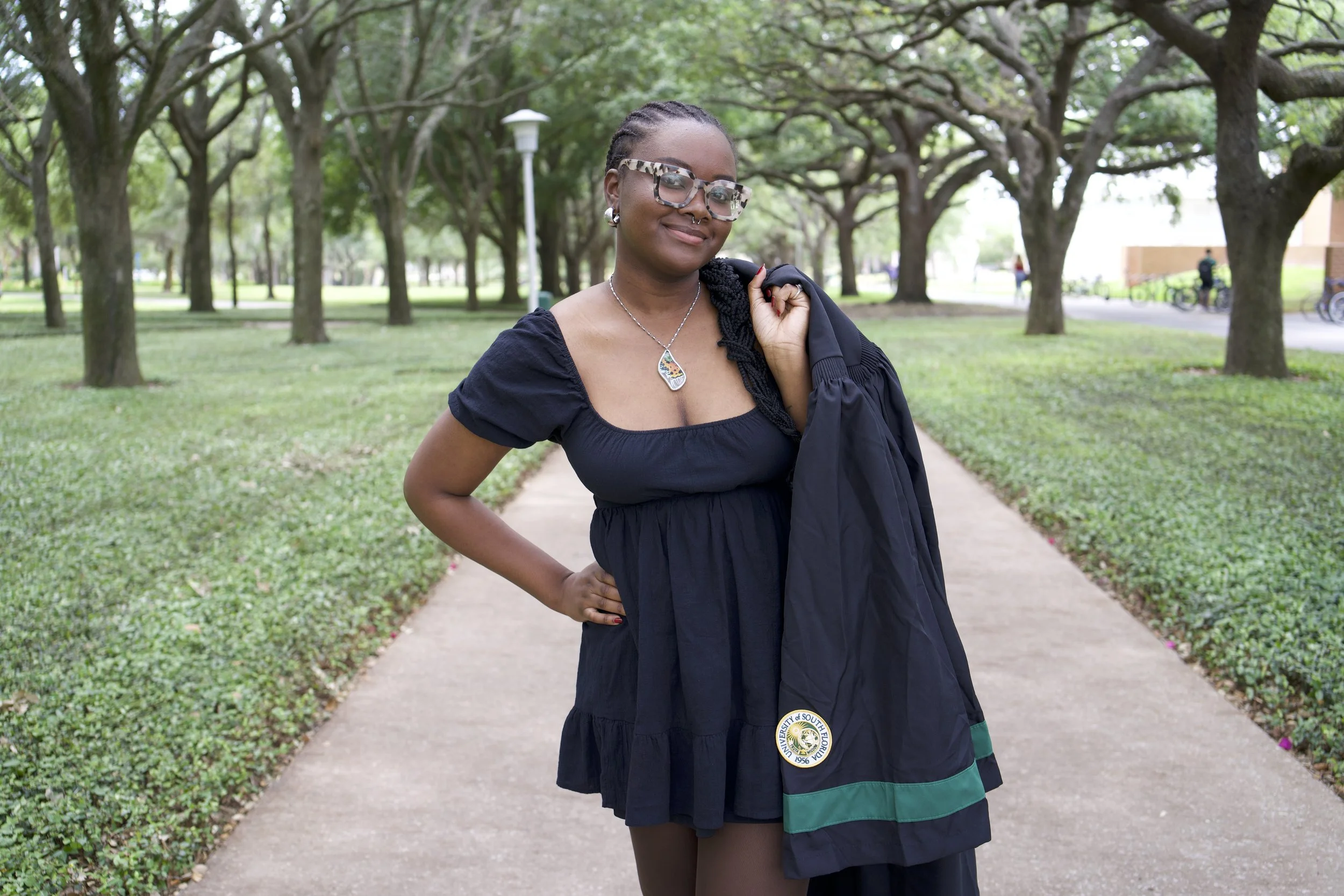 A woman in graduation attire standing on a pathway in a park with trees and greenery in the background, smiling and holding her jacket over her shoulder.