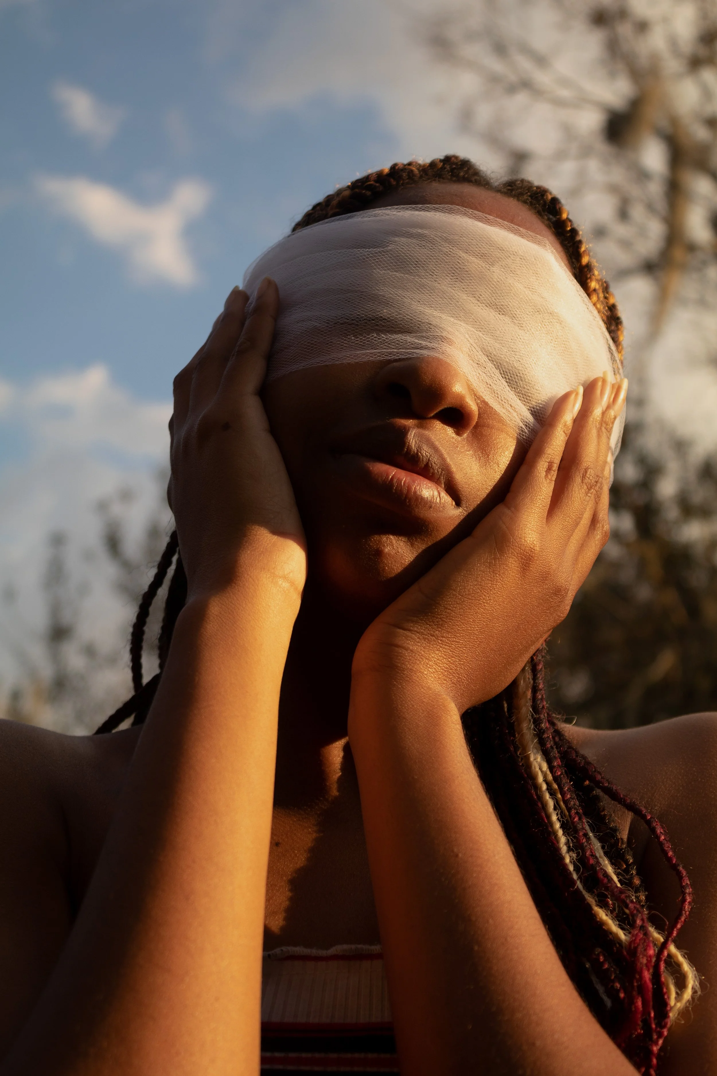 A woman outdoors with her eyes covered by a white cloth, touching her face with both hands, against a background of trees and a partly cloudy sky.