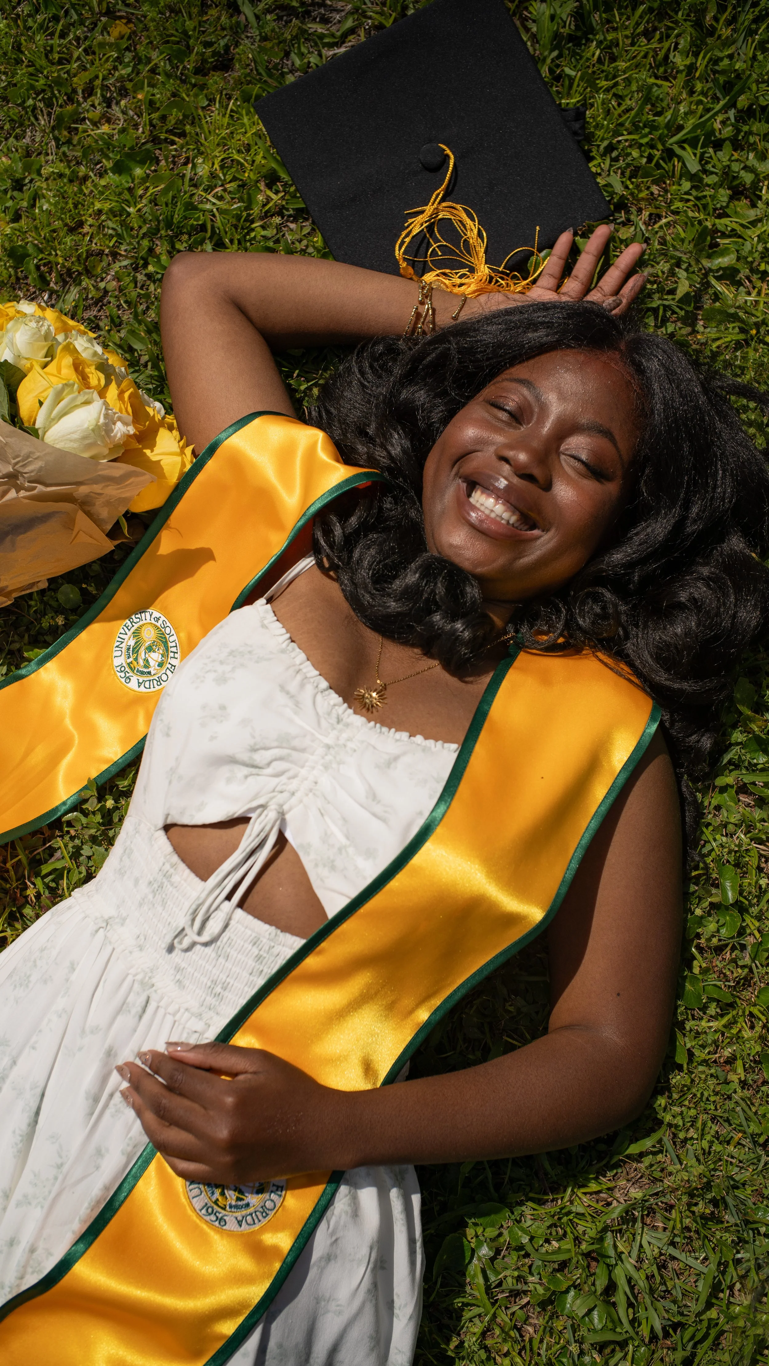 A woman lying on grass in her graduation cap and gown with a yellow and green stole, smiling with her eyes closed.