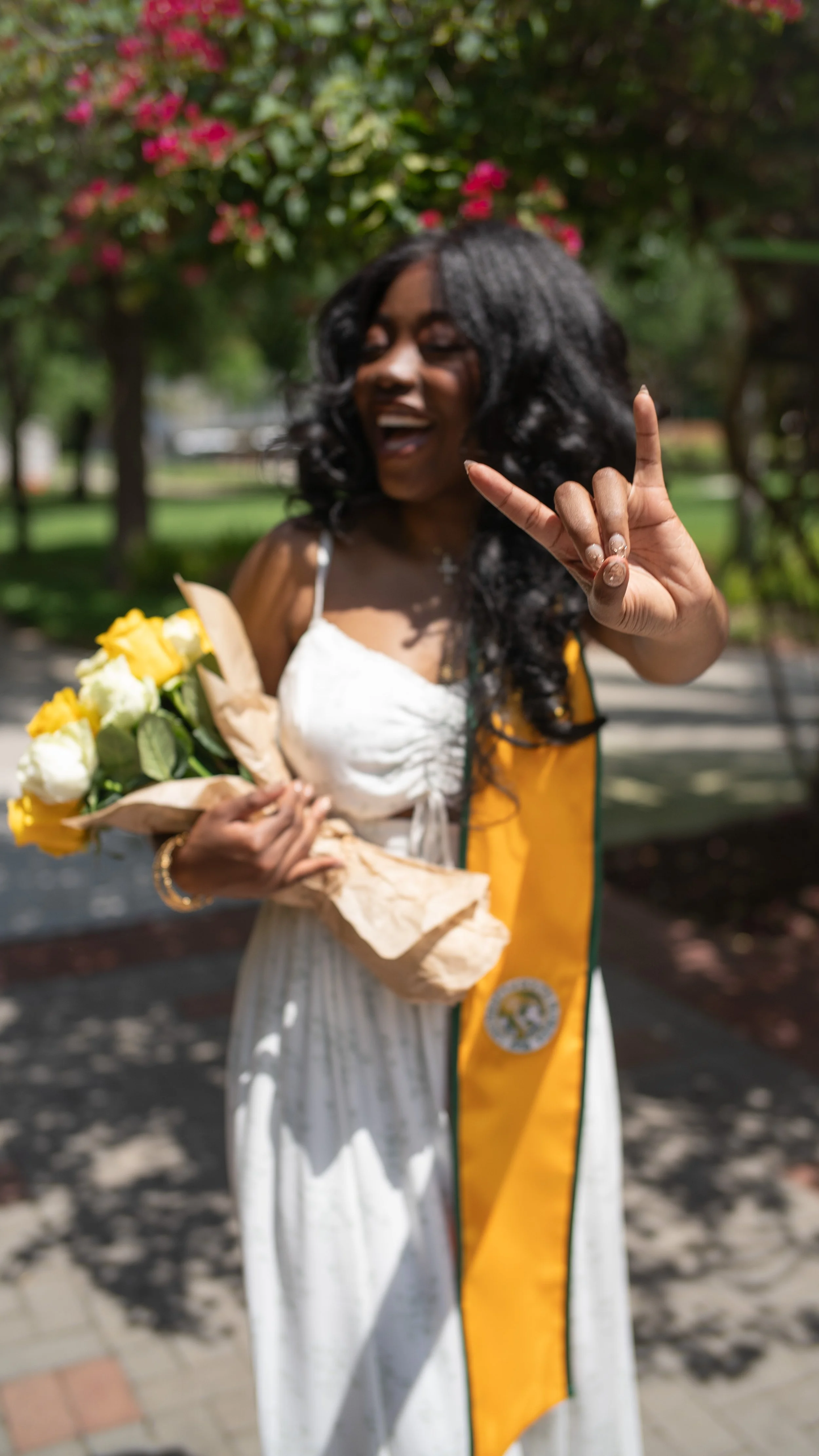 A woman in a white dress and yellow sash holding a bouquet of yellow and white roses, making a rock hand gesture and smiling outdoors with trees and pink flowers in the background.