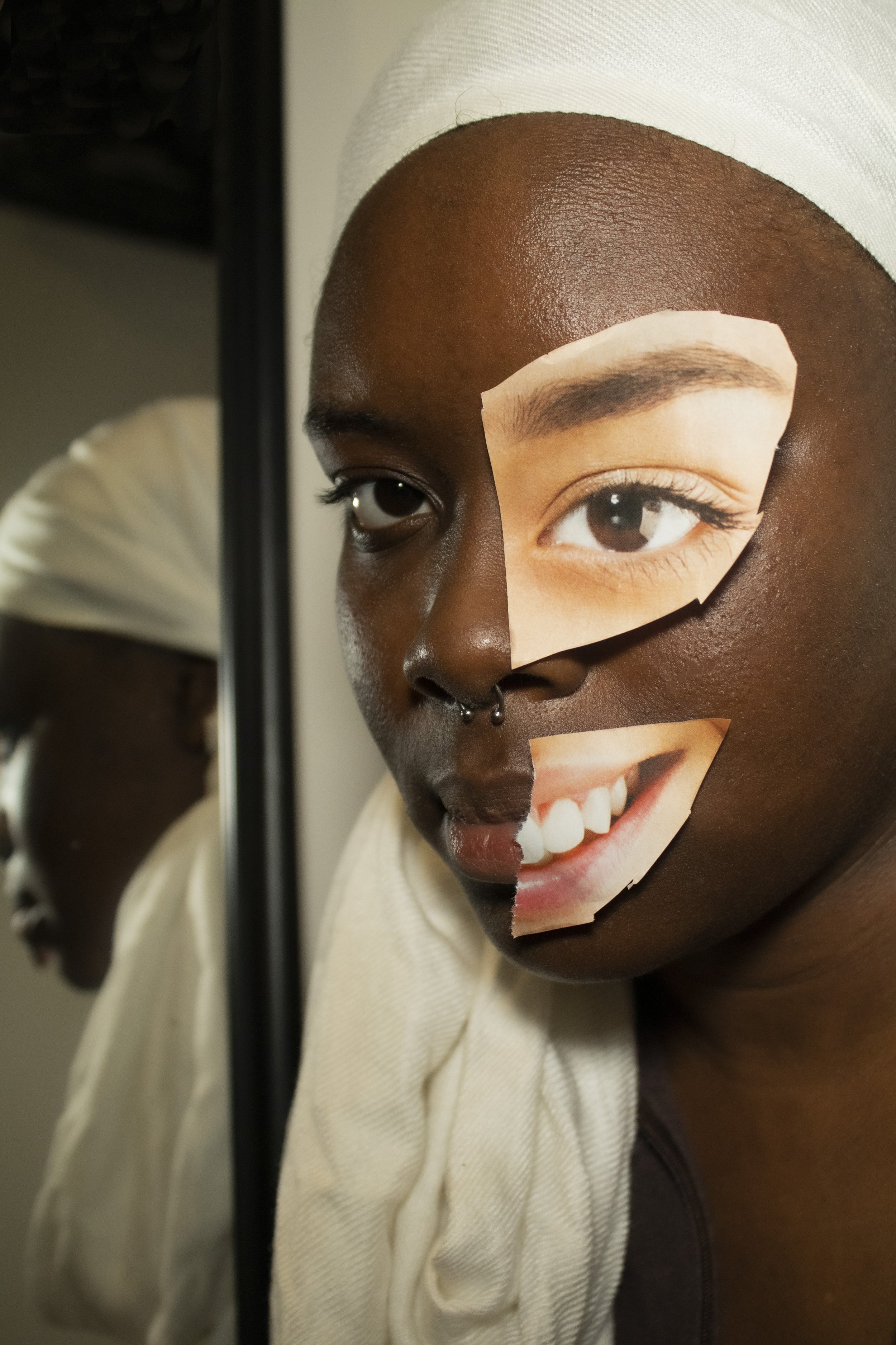 A person with dark skin and a septum piercing, looking in the mirror with a collage of cut-out photos of eyes and a smiling mouth on their face.