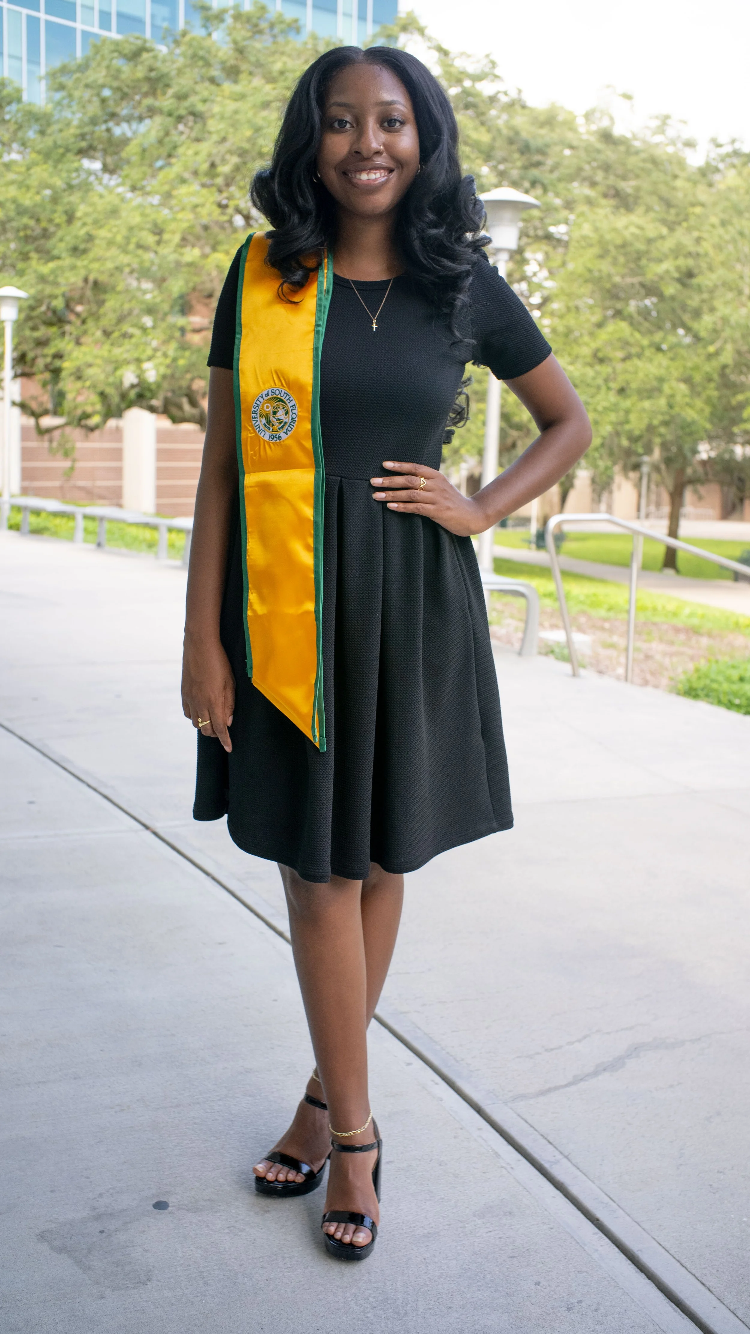 Young woman in a black dress and heels, standing outdoors with a yellow graduation stole, smiling at the camera.