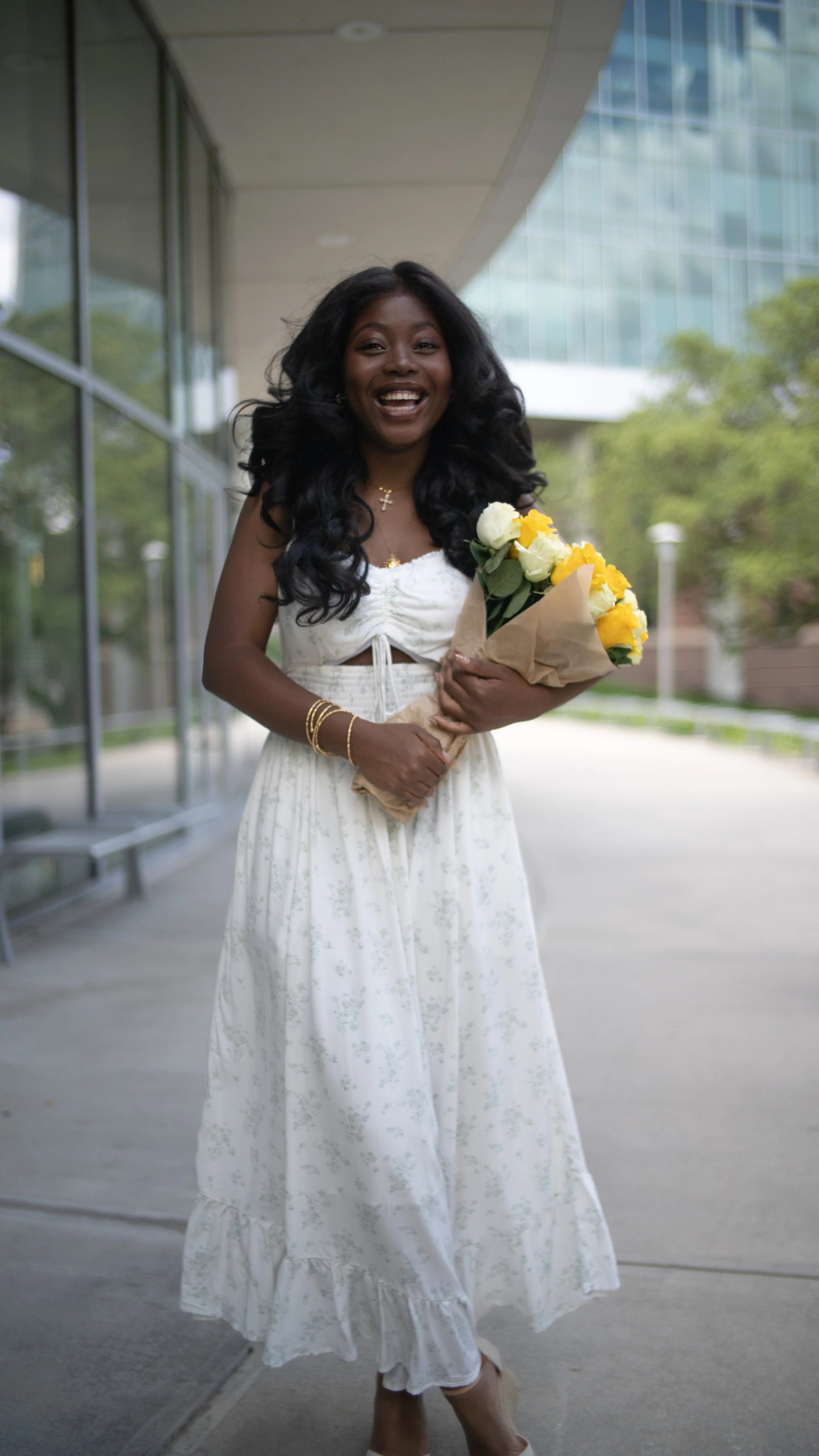 A woman with long black curly hair, wearing a white dress while holding a bouquet of yellow and white flowers, smiling outdoors near a modern building with glass windows.