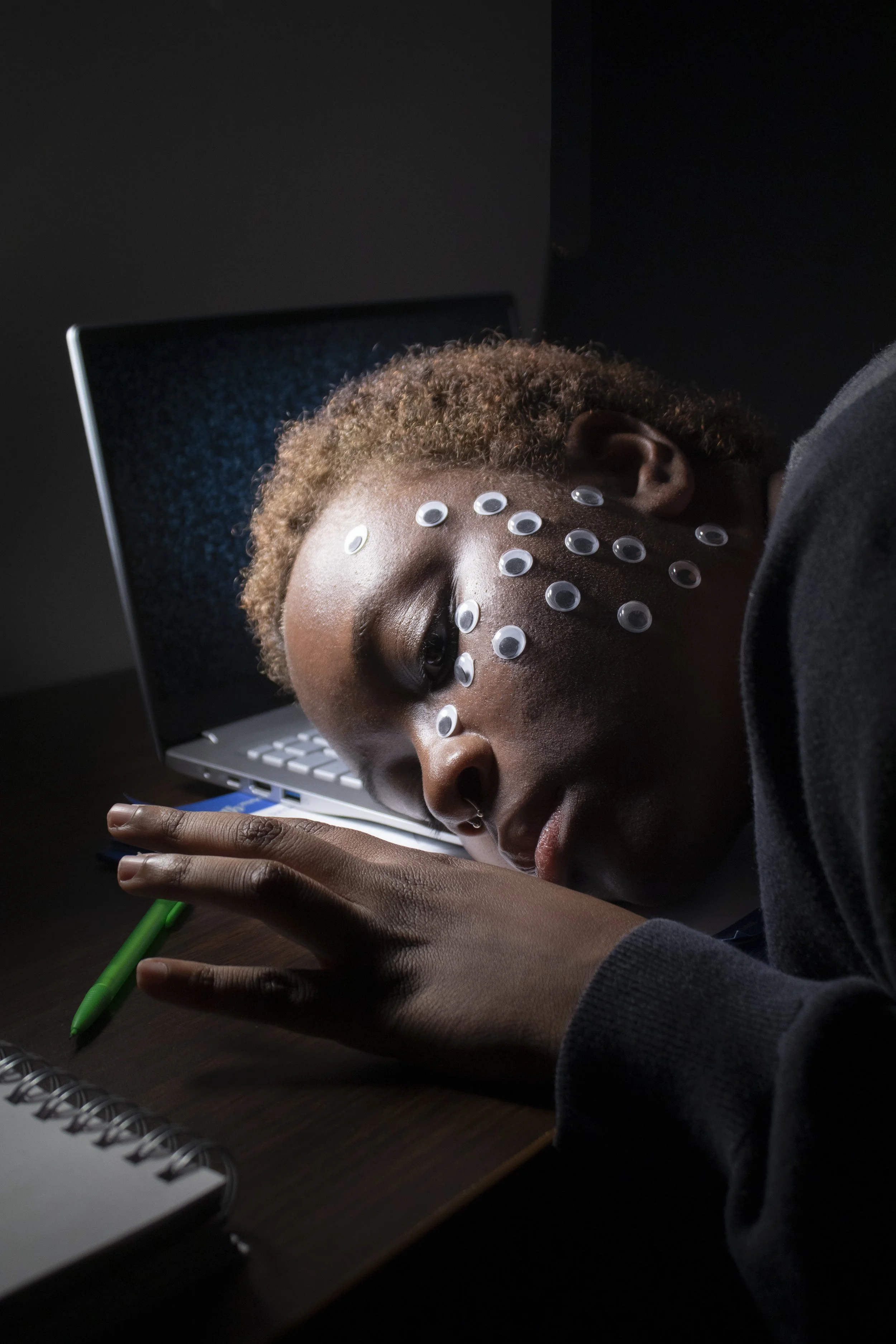 A person with short curly hair and eyelash extensions is lying face down on a desk, resting their head on their arm. They have multiple circular white goggles on their face, which could be for a skincare or therapy treatment. A laptop with a dark scr