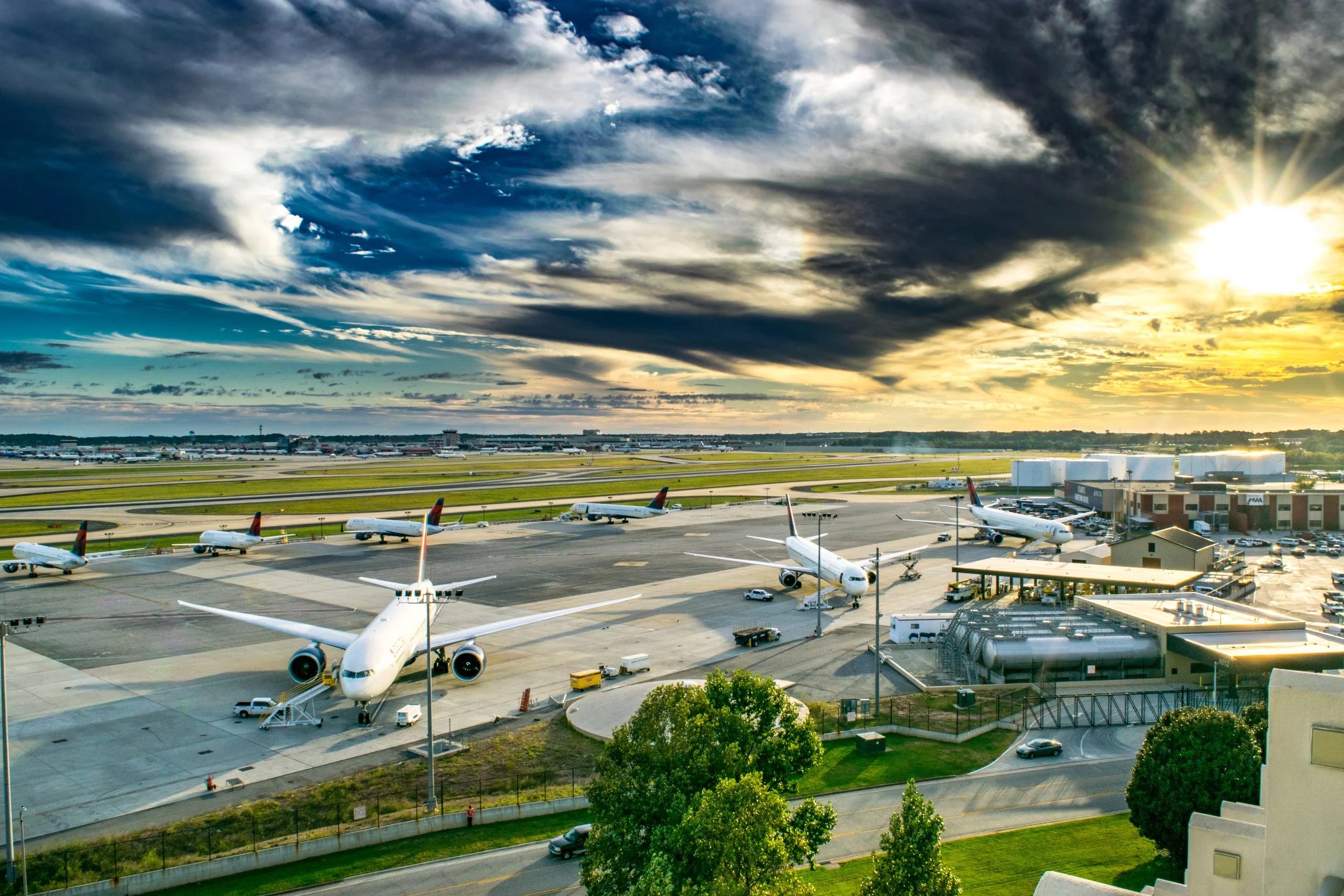 An airport tarmac with several parked airplanes, airport buildings, vehicles, and trees in the foreground, under a sky with dark and light clouds and the sun partially visible.