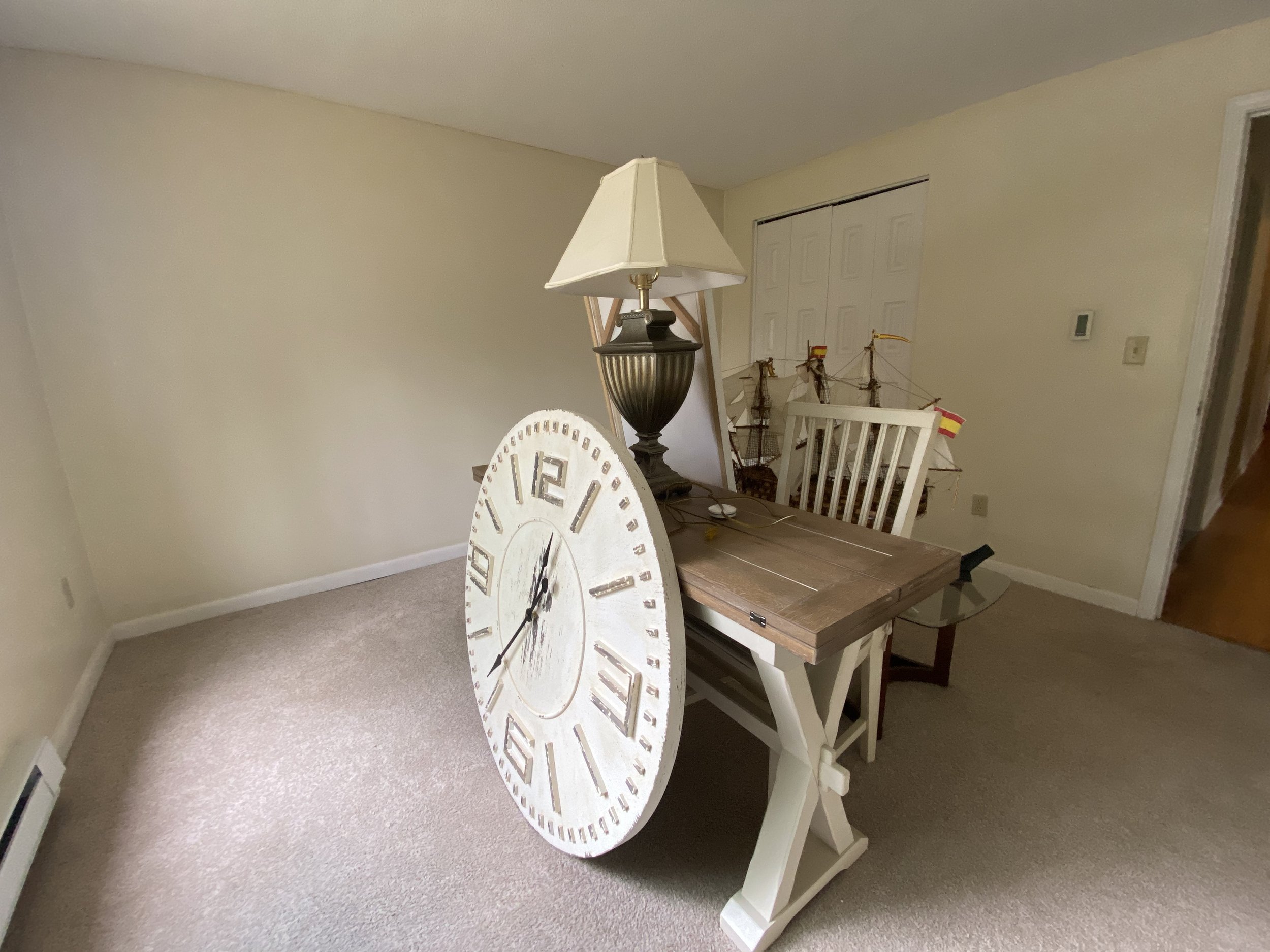 Empty room with a wooden table, a large wall clock leaning against the table, a table lamp, and a model ship in the background.