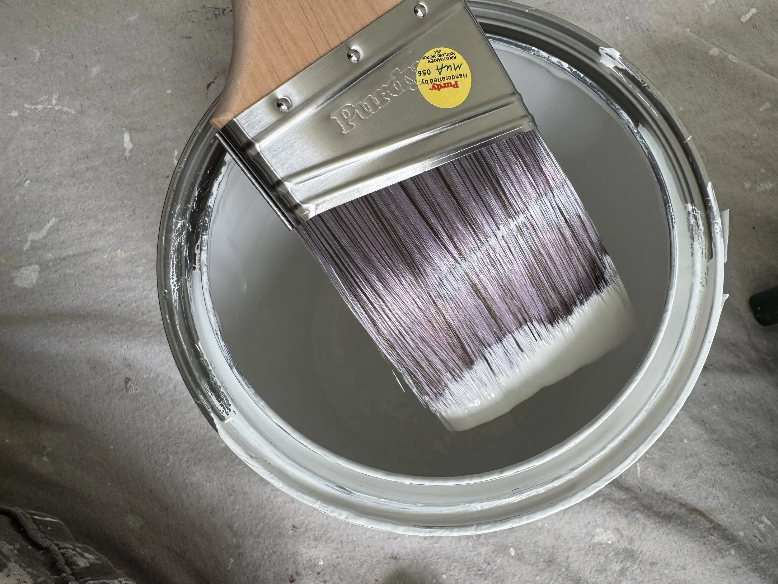 A paintbrush dipped in light gray paint above a paint bucket filled with the same light gray paint on a concrete floor.